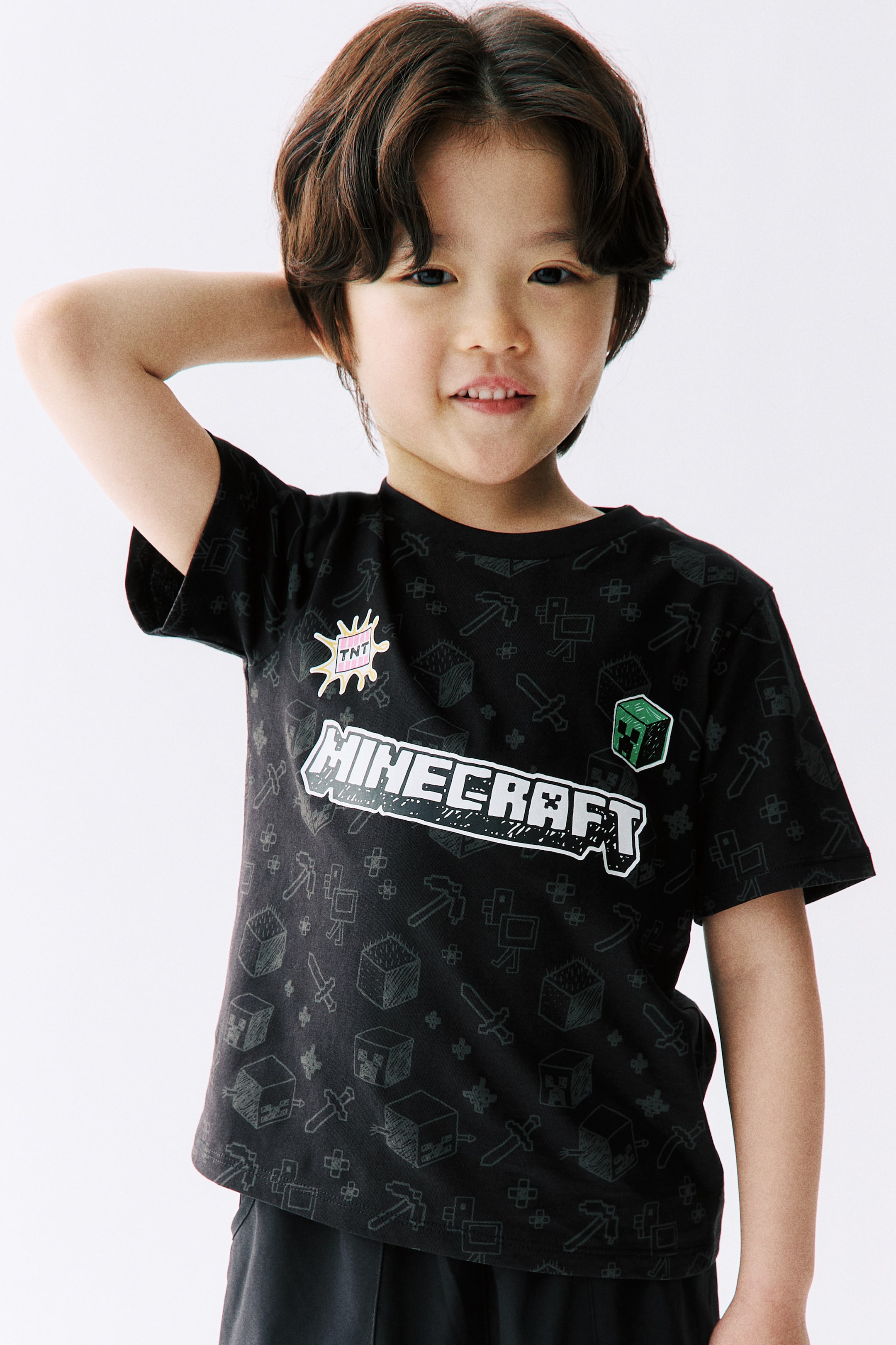 拡大画像を表示: 黒のTシャツには、さまざまなMinecraftアイコンのダークグレーのピクセルパターンが全面に表示されています。前面には、大きな白い「MINECRAFT」ロゴ、黄色とピンクの「TNT」爆発グラフィック、緑のクリーパーブロックグラフィックが特徴です。