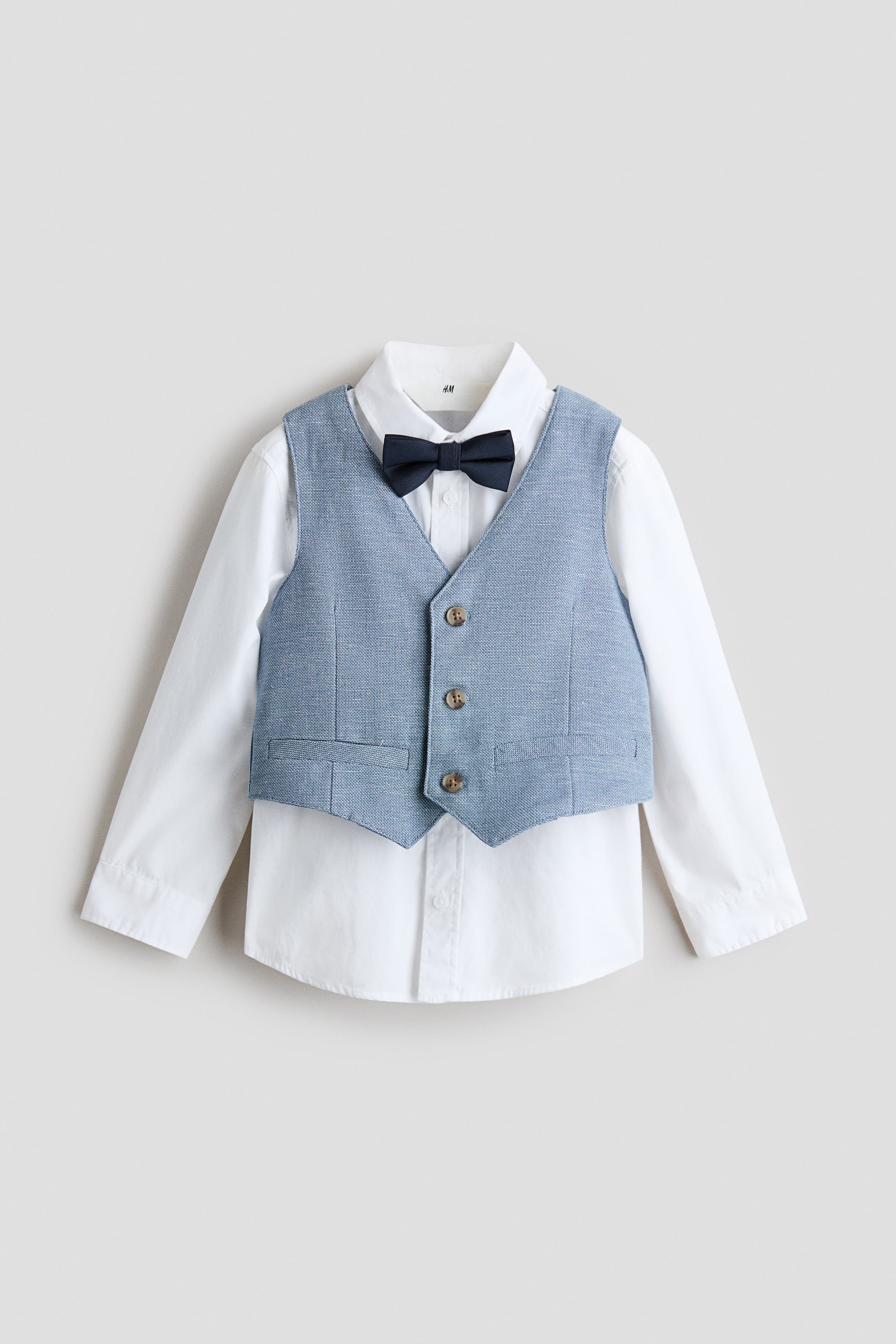 View larger image: This formal ensemble features a white long-sleeve shirt with a collar and buttoned cuffs, layered with a light blue woven vest adorned with three brown buttons and faux pockets. A dark blue pre-tied bow tie is neatly positioned at the collar, all displayed in a front-facing view.