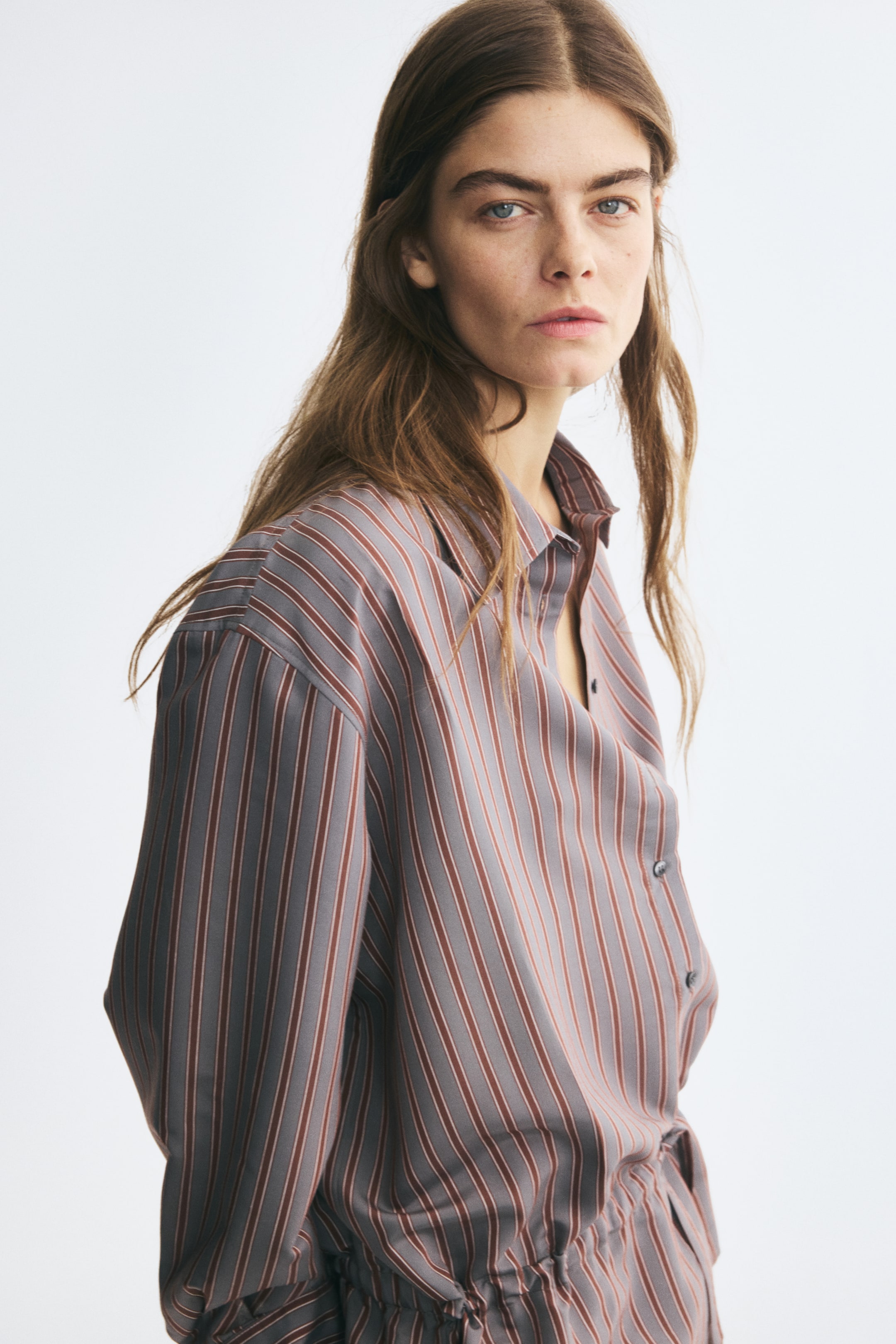 Ver imagen más grande: La modelo lleva una camisa de manga larga a rayas con rayas verticales grises y marrones. La camisa se presenta con cuello abierto y se combina con un pantalón a rayas a juego.