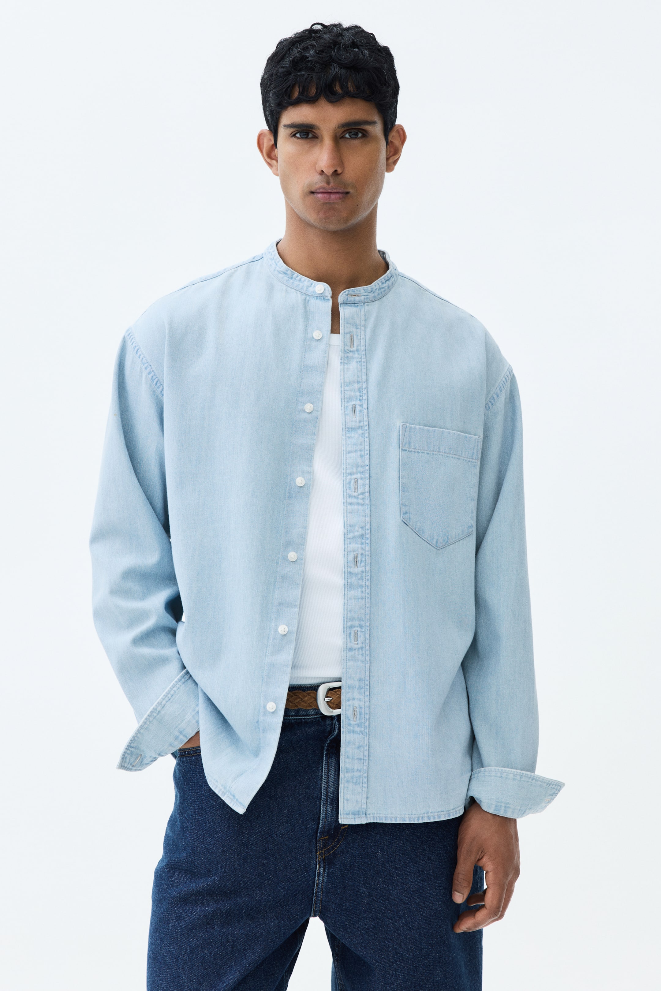 View larger image: The model is seen wearing a light blue rigid cotton denim shirt featuring a band collar and a classic front design. It includes a patch chest pocket for added style. The shirt is styled with long sleeves that have rolled buttoned cuffs and a gently rounded hem. It is worn open over a white undershirt, offering a casual yet refined look.