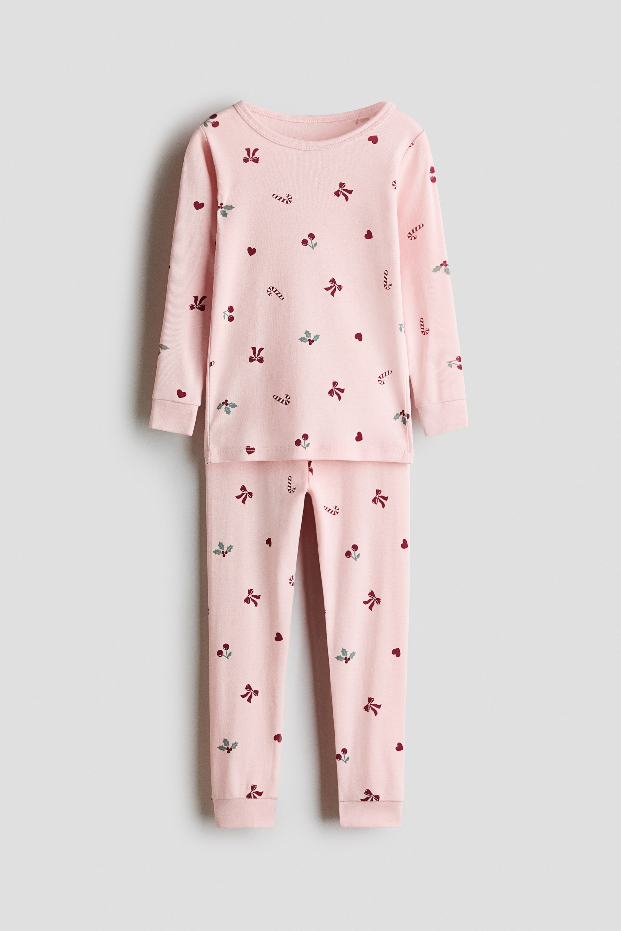 View larger image: A light pink cotton jersey pajama set is shown from the front, featuring an all-over festive print of red bows, candy canes, hearts, and holly. The long-sleeved top has a round neckline and ribbed cuffs, paired with matching pants that feature an elasticated waist and ribbed cuffed hems.