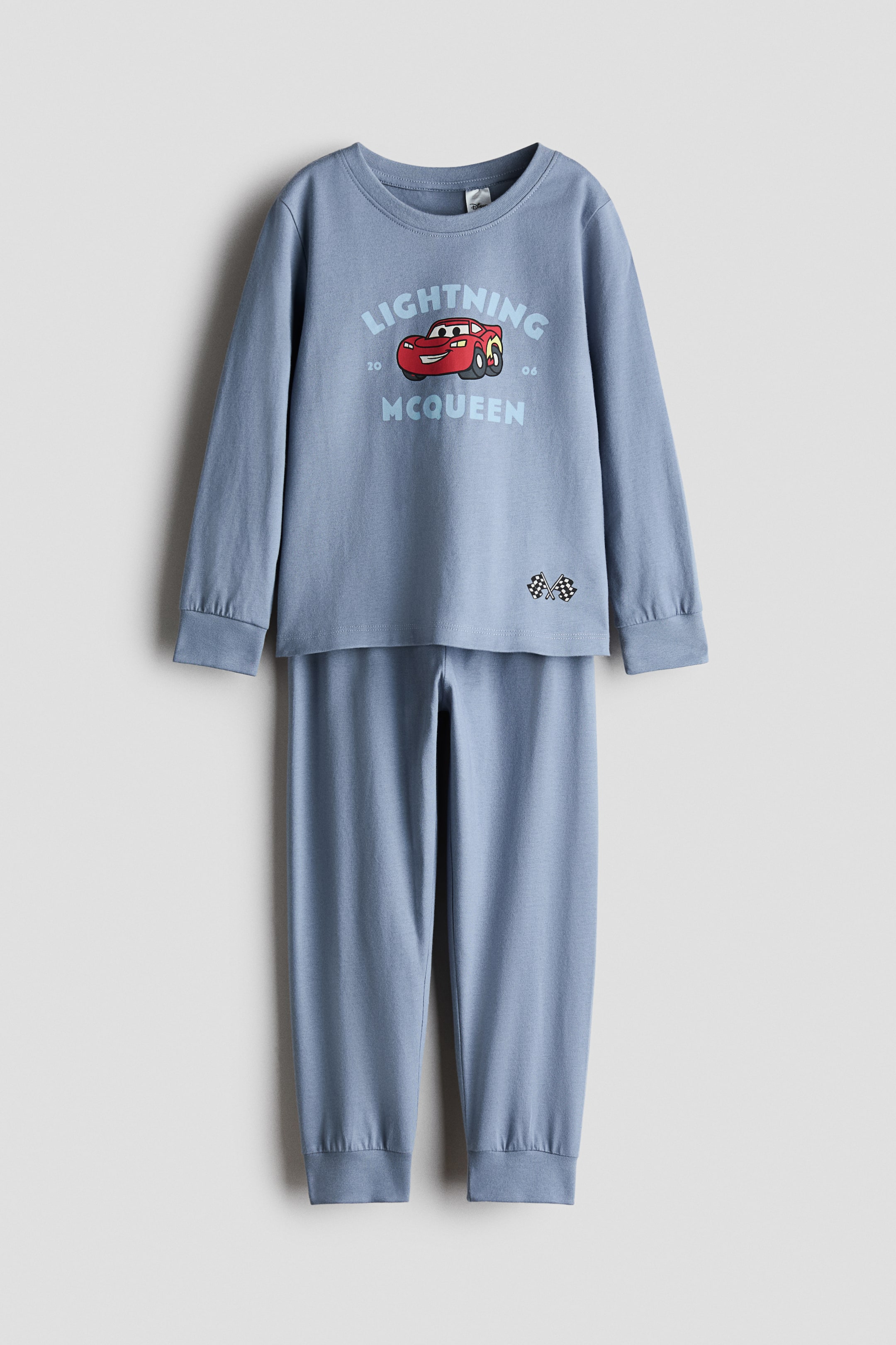 Büyük resmi görüntüle: Açık mavi pijama takımı, yumuşak baskılı pamuklu jarse kumaştan yapılmıştır ve düz olarak asılı durmaktadır. Uzun kollu üst kısmı, dar bir şerit ile yuvarlak bir yaka ve "Lightning McQueen" grafik tasarımıyla kırmızı bir yarış arabası ve damalı bayraklar içermektedir. Uyumlu alt kısmı ise lastikli bir bel ve nervürlü paçalar göstermektedir.