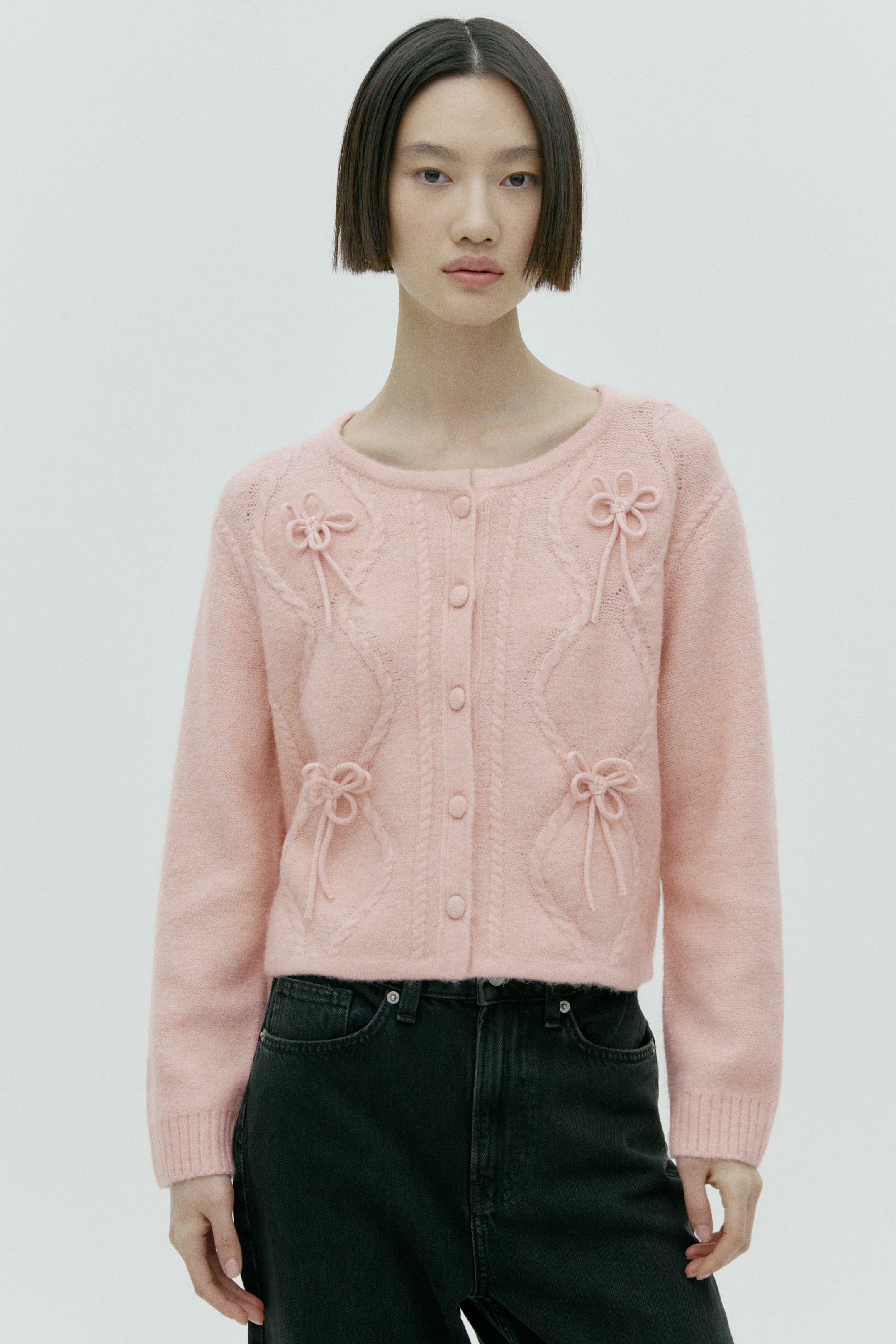 이미지 확대 보기: 핑크 카디건은 라운드 네크라인과 동일한 핑크 버튼의 앞면 버튼 여밈을 특징으로 합니다. 앞면에는 다이아몬드 모양을 이루는 케이블 니트 패턴이 들어가며, 여섯 개의 입체적인 리본 장식이 더해져 있습니다. 또한 골지 처리된 소맷단의 긴 소매와 크롭 기장의 골지 밑단을 갖추고 있습니다.
