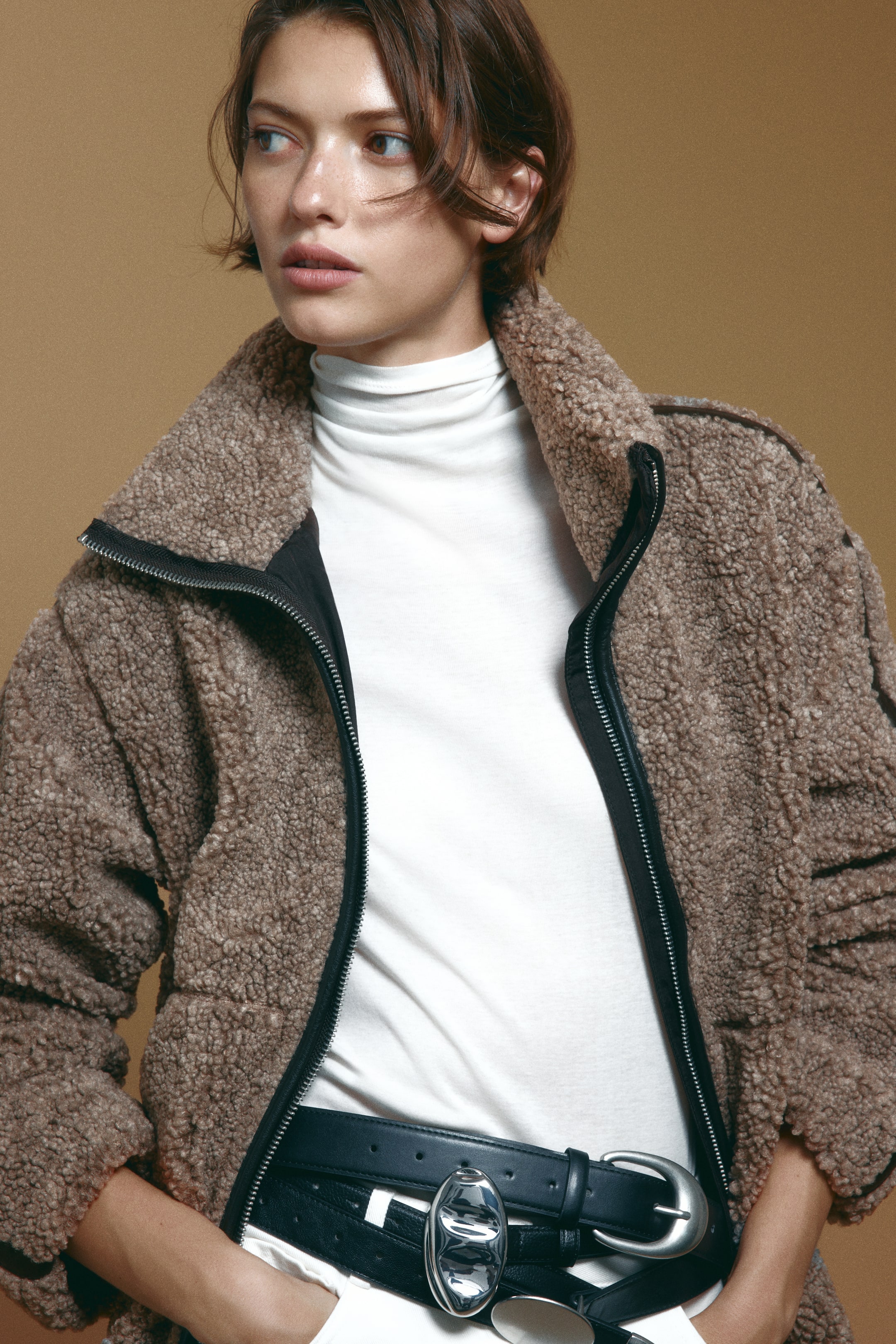 Größeres Bild anzeigen: Ein oversized geschnittener brauner Teddyjacke mit weitem Stehkragen und schwarzer beschichteter Stoffkante entlang des Frontreißverschlusses wird vom Model offen getragen. Die Jacke ist über einem weißen Rollkragentop und weißen Hosen getragen, die mit einem schwarzen Ledergürtel gestylt sind.