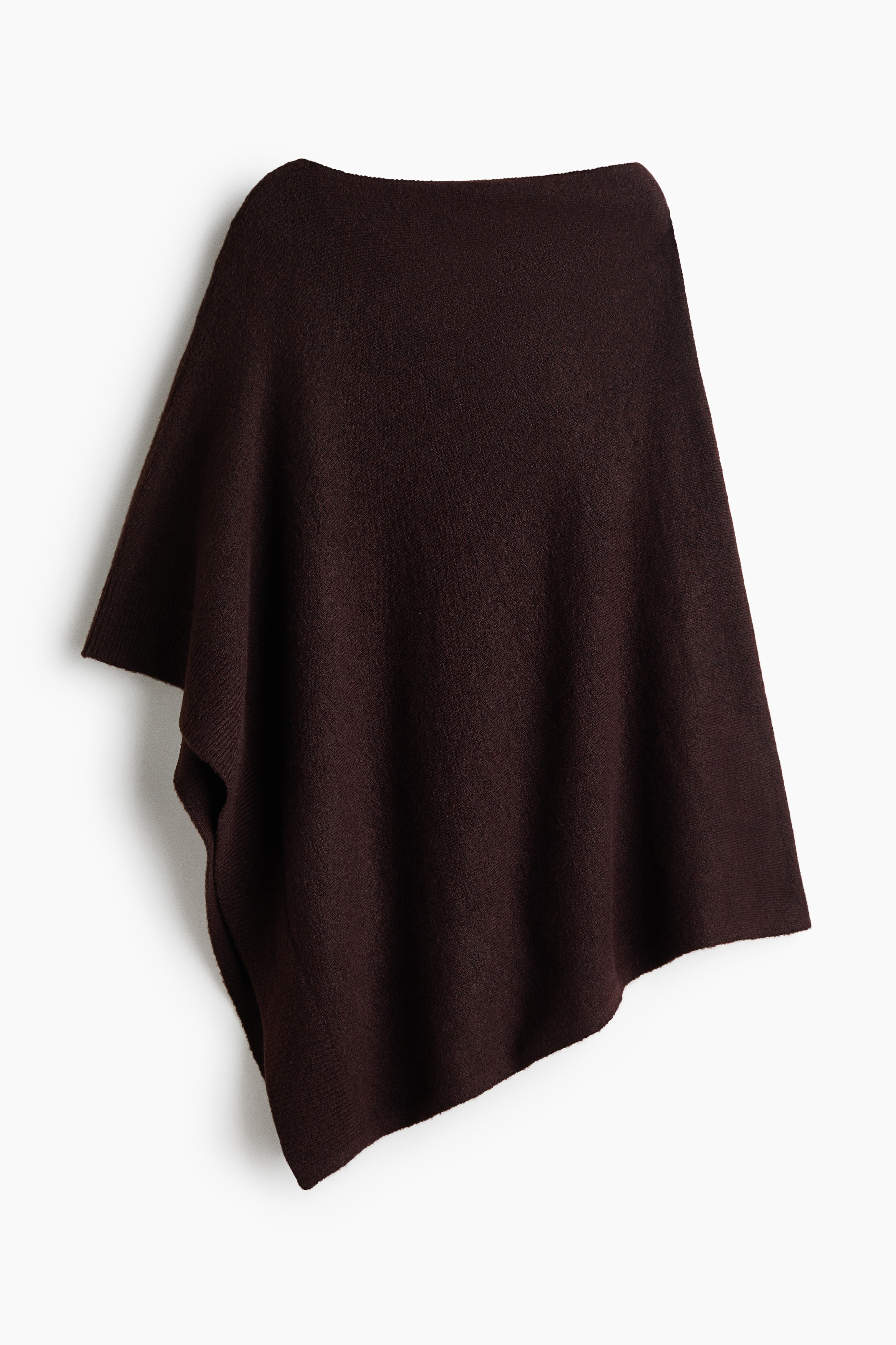 Een oversized, asymmetrische cape in donkerbruin fijn gebreid materiaal wordt door het model gedragen over een witte broek. De cape heeft een hoge boothals en valt losjes over de torso met een onregelmatige, puntige zoomlijn.