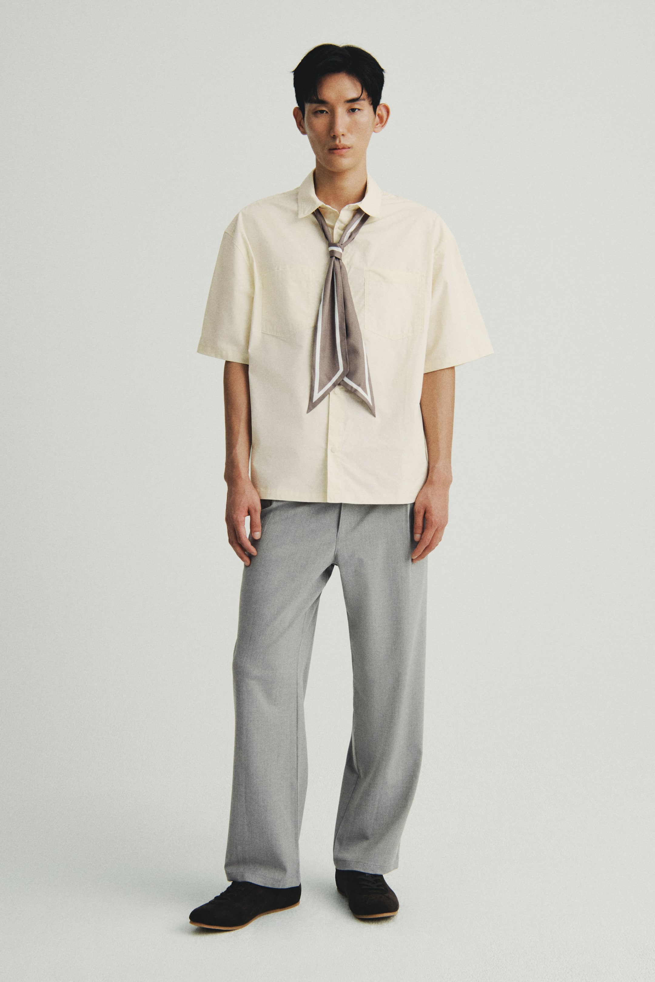 Xem ảnh lớn hơn: Một chiếc áo sơ mi màu sáng, tay ngắn đang được người mẫu mặc, có cổ áo gấp và hai túi ngực đắp. Một chiếc khăn hoa văn được buộc quanh cổ áo, tôn lên dáng áo rộng và gấu áo cắt thẳng.