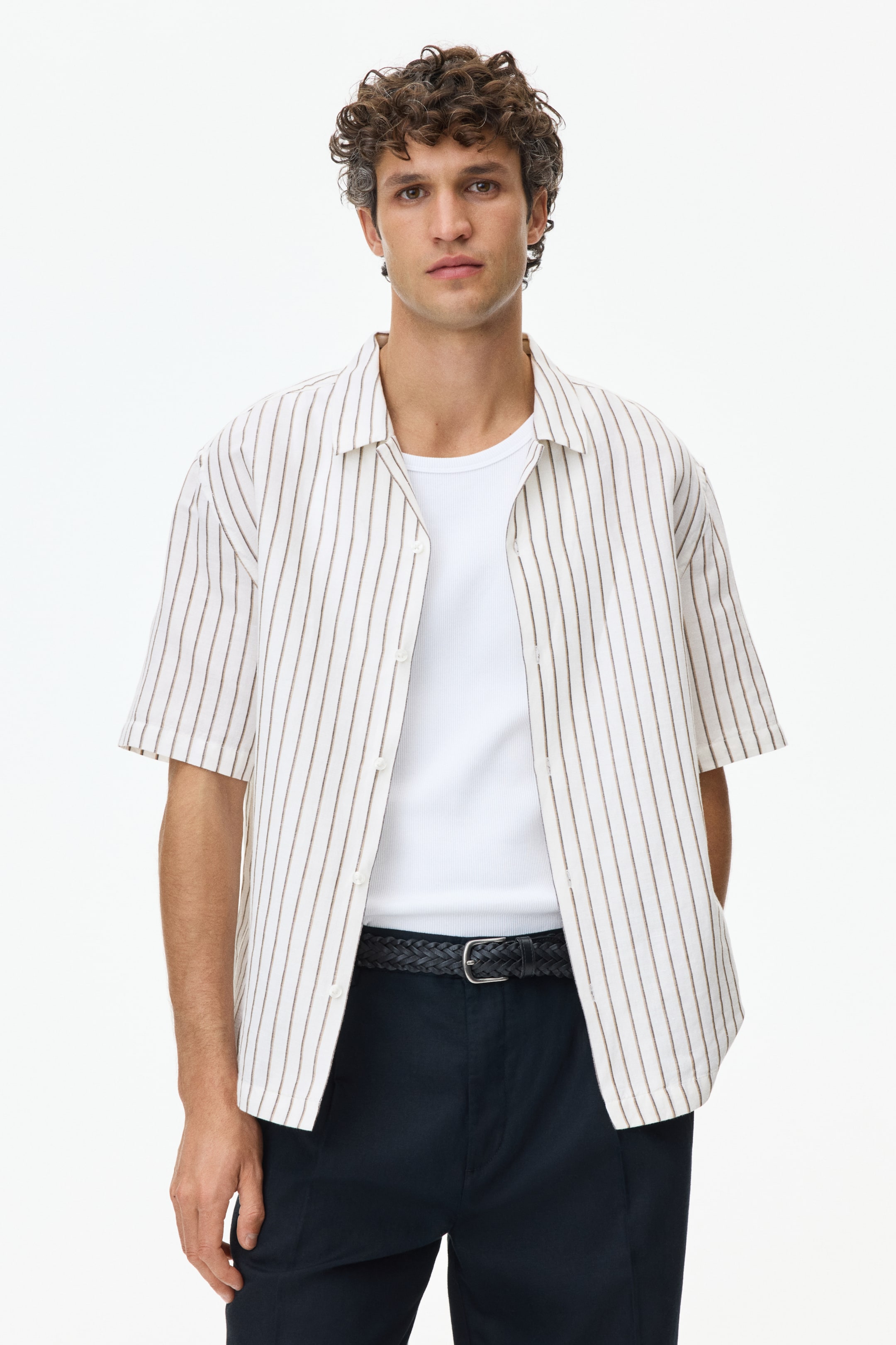 Agrandir l'image: La chemise blanche à manches courtes avec de fines rayures verticales marron est présentée de face, portée ouverte sur un débardeur blanc côtelé. Elle est dotée d'un col évasé et de boutons blancs visibles le long de la patte de boutonnage, atteignant juste en dessous de la taille et révélant un pantalon foncé avec une ceinture tressée foncée.