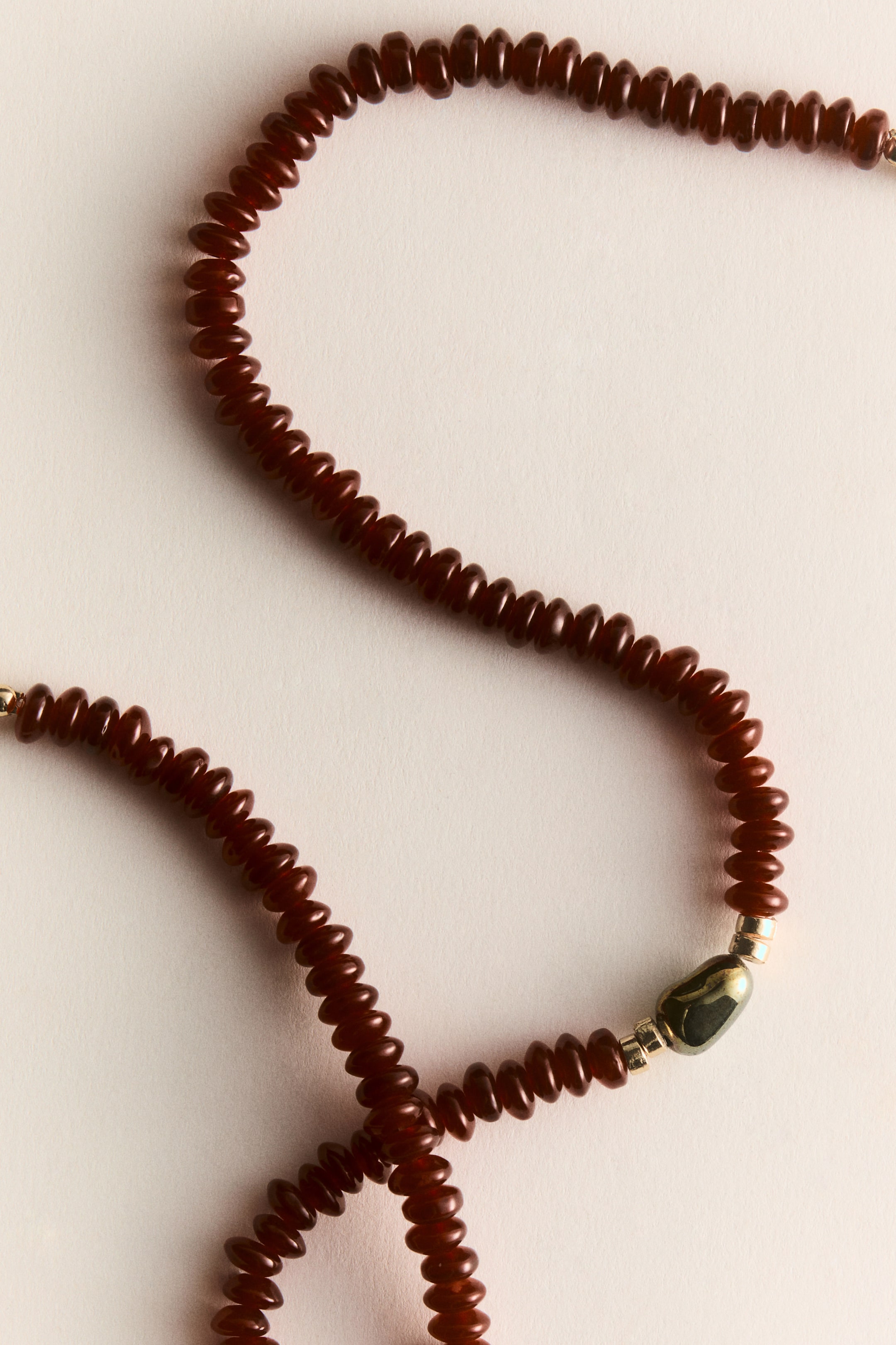 View larger image: The necklace is elegantly arranged in an S-curve, highlighting a string of numerous dark reddish-brown, flattened disc-shaped beads. At the center of the strand, there's an irregularly shaped dark bead adorned with metallic flecks, flanked by two small gold-colored spacer beads.
