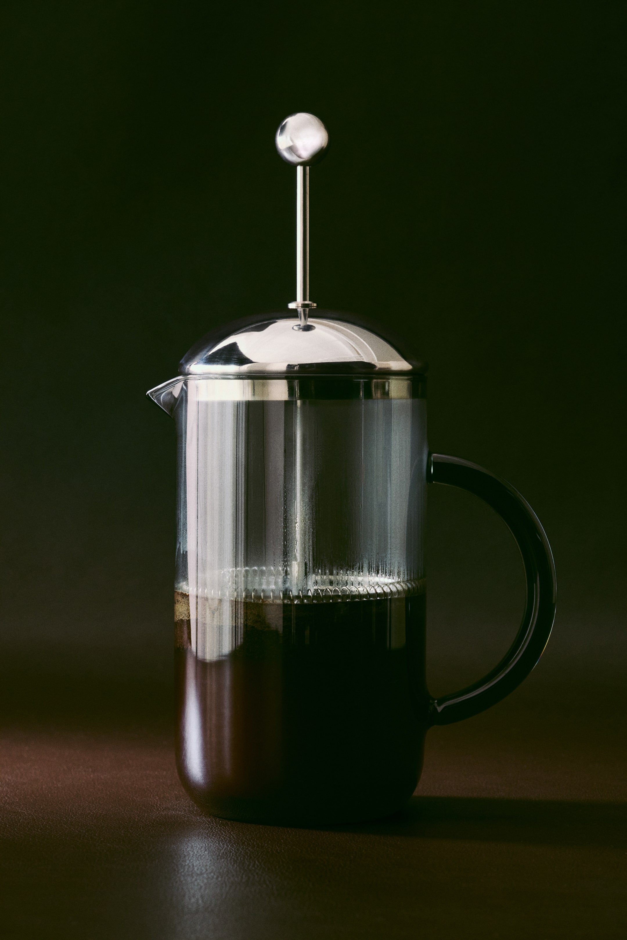 Agrandir l'image: Une cafetière à piston en verre borosilicaté transparent est présentée à la verticale, contenant du café infusé foncé dans sa moitié inférieure. Elle est dotée d’un couvercle en acier inoxydable en forme de dôme, avec un piston allongé surmonté d’un bouton sphérique, ainsi que d’une poignée arrondie sombre à droite et d’un bec verseur à gauche.