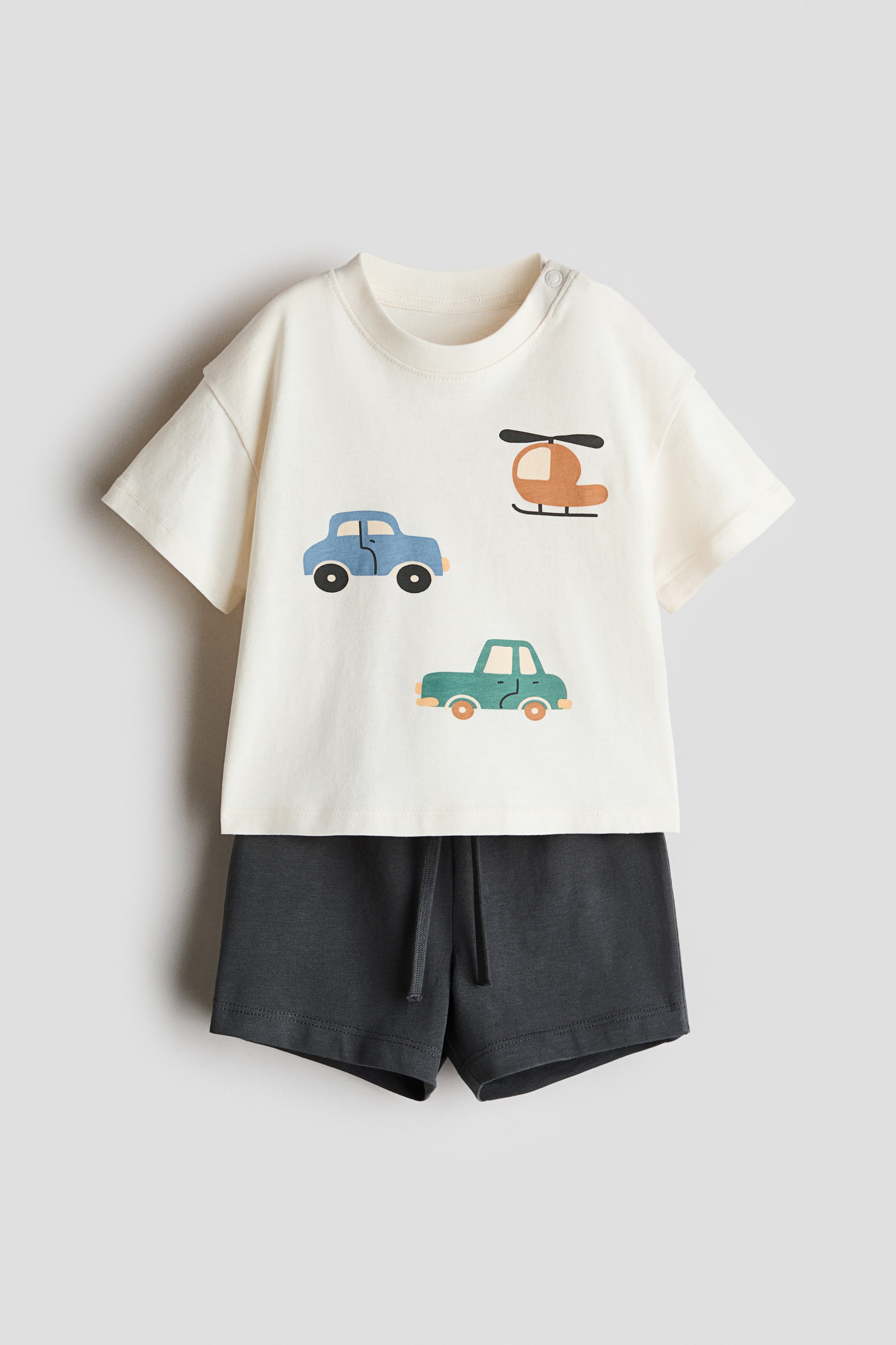 View larger image: Introducing a 2-piece cotton set showcased from the front. The off-white short-sleeved t-shirt boasts a crew neck with two snap buttons on the left shoulder, adorned with cartoon vehicles. Complementing the set are dark grey shorts featuring a visible drawstring waist.