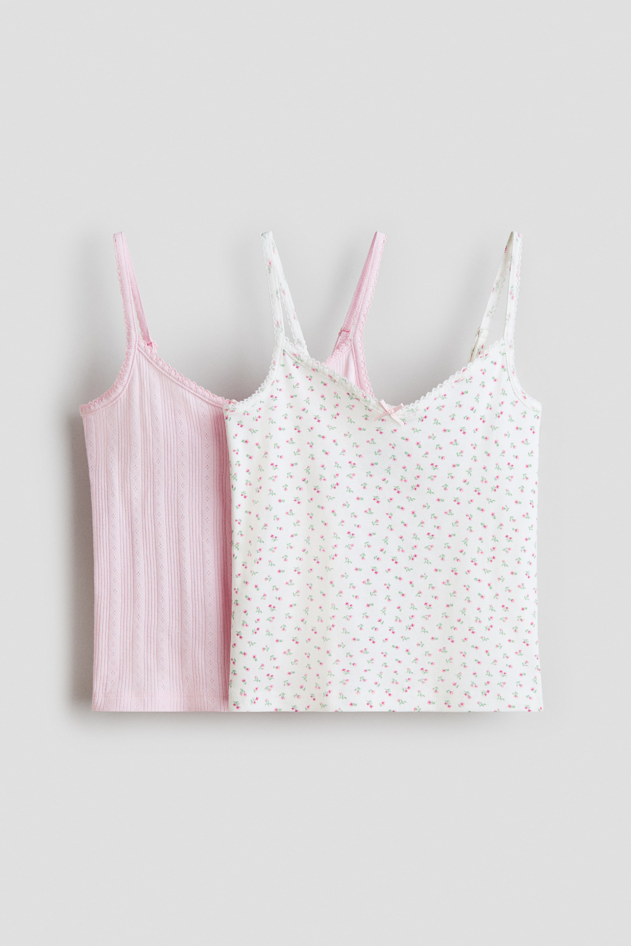 Visa större bild: Ett set med två linnen ligger platt, med det rosa linnet delvis synligt bakom det vita blommiga linnet. Det rosa linnet har en ribbstickning med hålsömsdetaljer, medan det vita linnet visar ett litet rosa blommönster och en liten rosa rosett vid den främre halsringningen, båda avslutade med en delikat spetskant längs halsringningen och axelbanden.