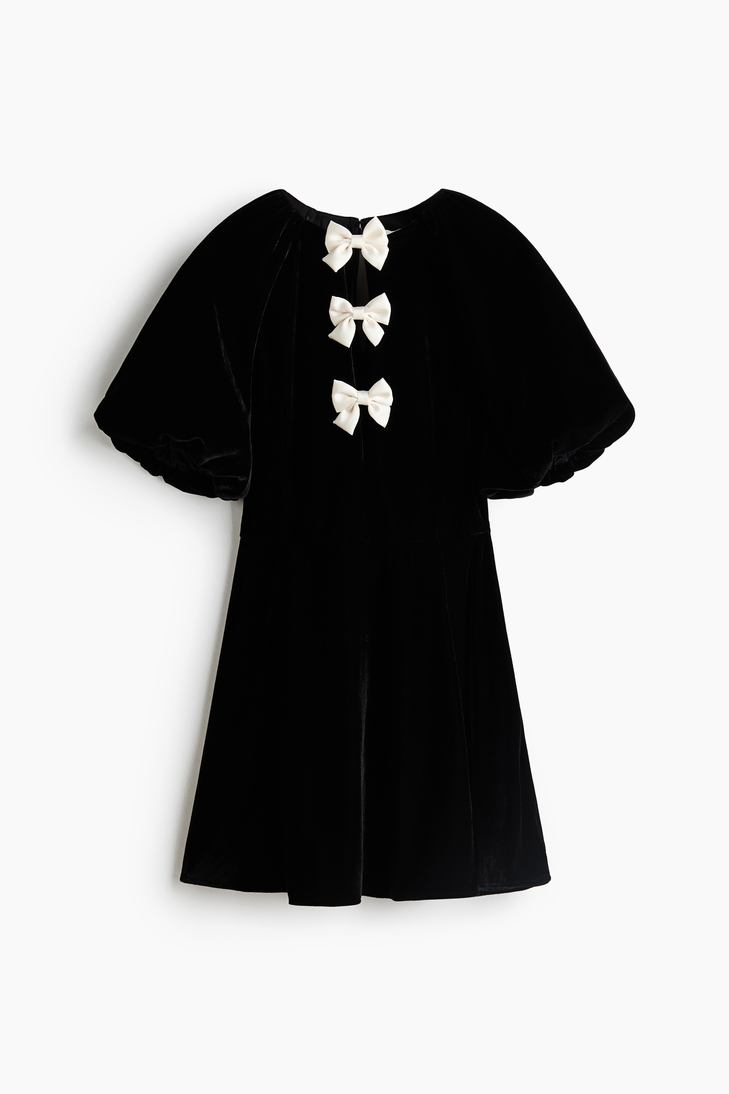 The model is wearing a short black velvet dress featuring elbow-length puff sleeves, a fitted bodice and a flared skirt. The dress has a deep front V-neckline detailed with three decorative white bows, which the model is lightly touching with one hand.