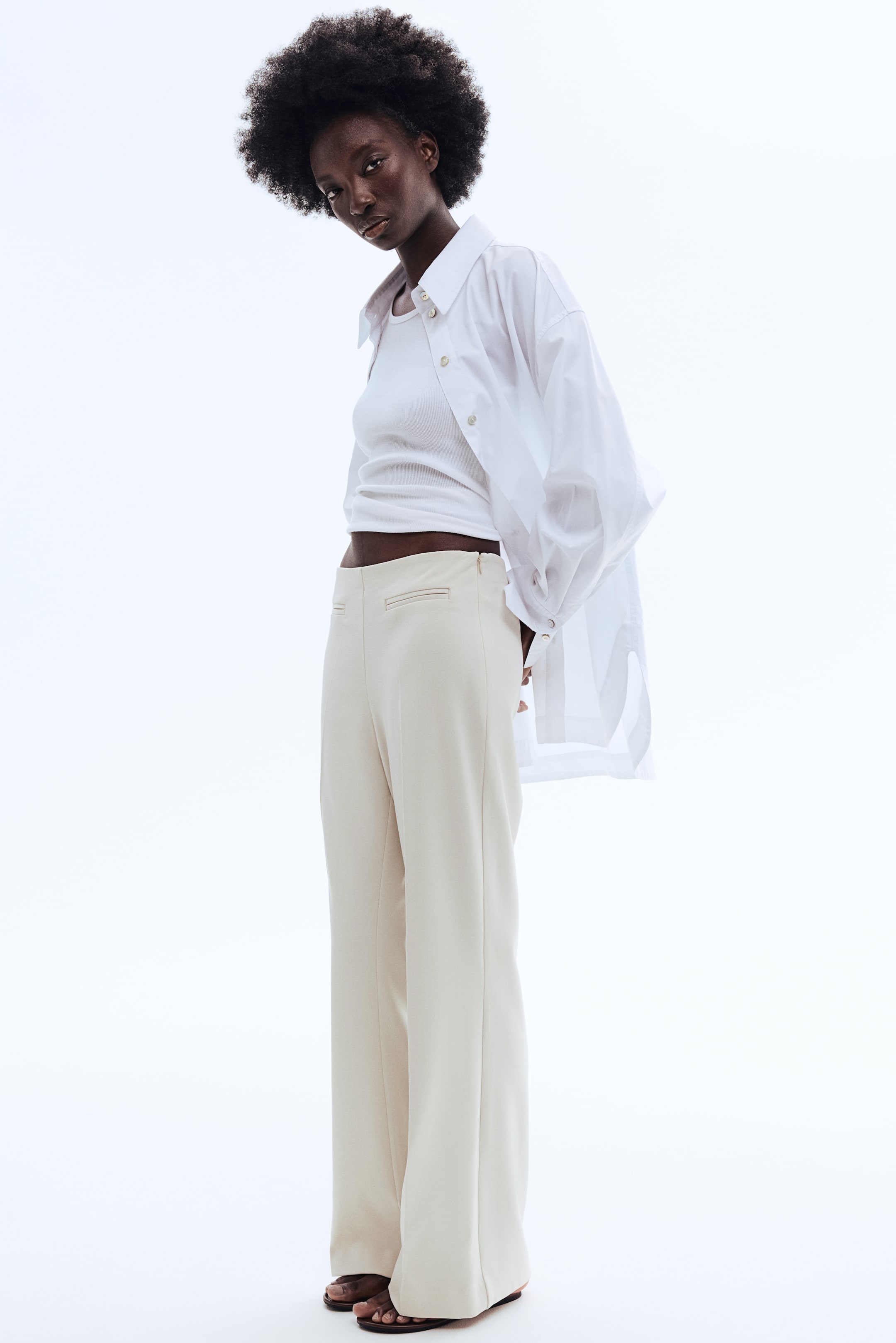 View larger image: Cream-coloured high-waisted wide-leg trousers are showcased from a slight front-left angle. They feature a straight fit with crease lines down the front of the legs, a visible side zipper closure at the left hip, and welt pockets at the back waistband. Styled with a white tank top and an oversized white shirt, these trousers offer a chic and versatile look.