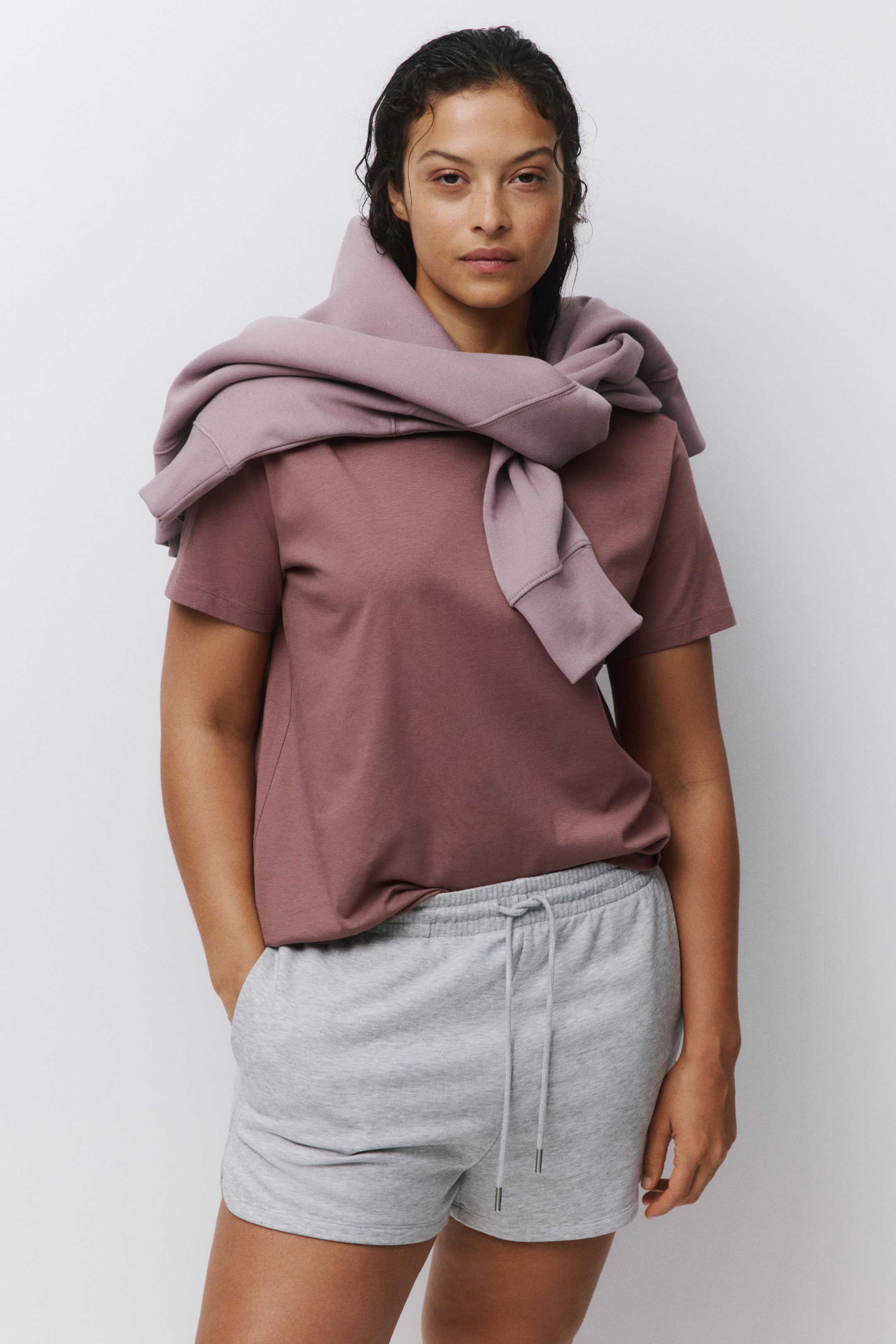 Преглед на по-голямо изображение: Тъмнорозова тениска с къс ръкав се носи с къси панталони в светлосиво, с връзка на талията и метални накрайници. Лилав суитшърт е преметнат през раменете, с ръкавите завързани отпред, показващи оребрени маншети.