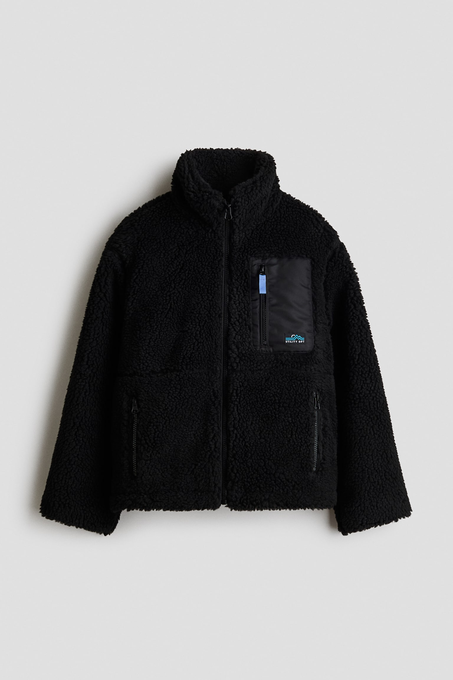 Une veste en molleton teddy noire est prÃ©sentÃ©e de face, mettant en valeur un col montant haut, une fermeture frontale Ã  glissiÃ¨re intÃ©grale et des Ã©paules lÃ©gÃ¨rement tombantes. Elle prÃ©sente une poche poitrine verticale zippÃ©e en tissu tissÃ© noir lisse sur le cÃ´tÃ© gauche, avec un petit Ã©cusson graphique, ainsi que deux poches latÃ©rales zippÃ©es.