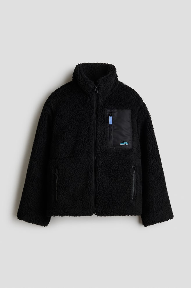 A black teddy fleece jacket is facing front, showcasing a high stand collar, a full-zip front closure, and gently dropped shoulders. It is featuring a vertical zipped chest pocket in smooth woven black fabric on the left side with a small graphic patch, along with two zipped side pockets. - 1