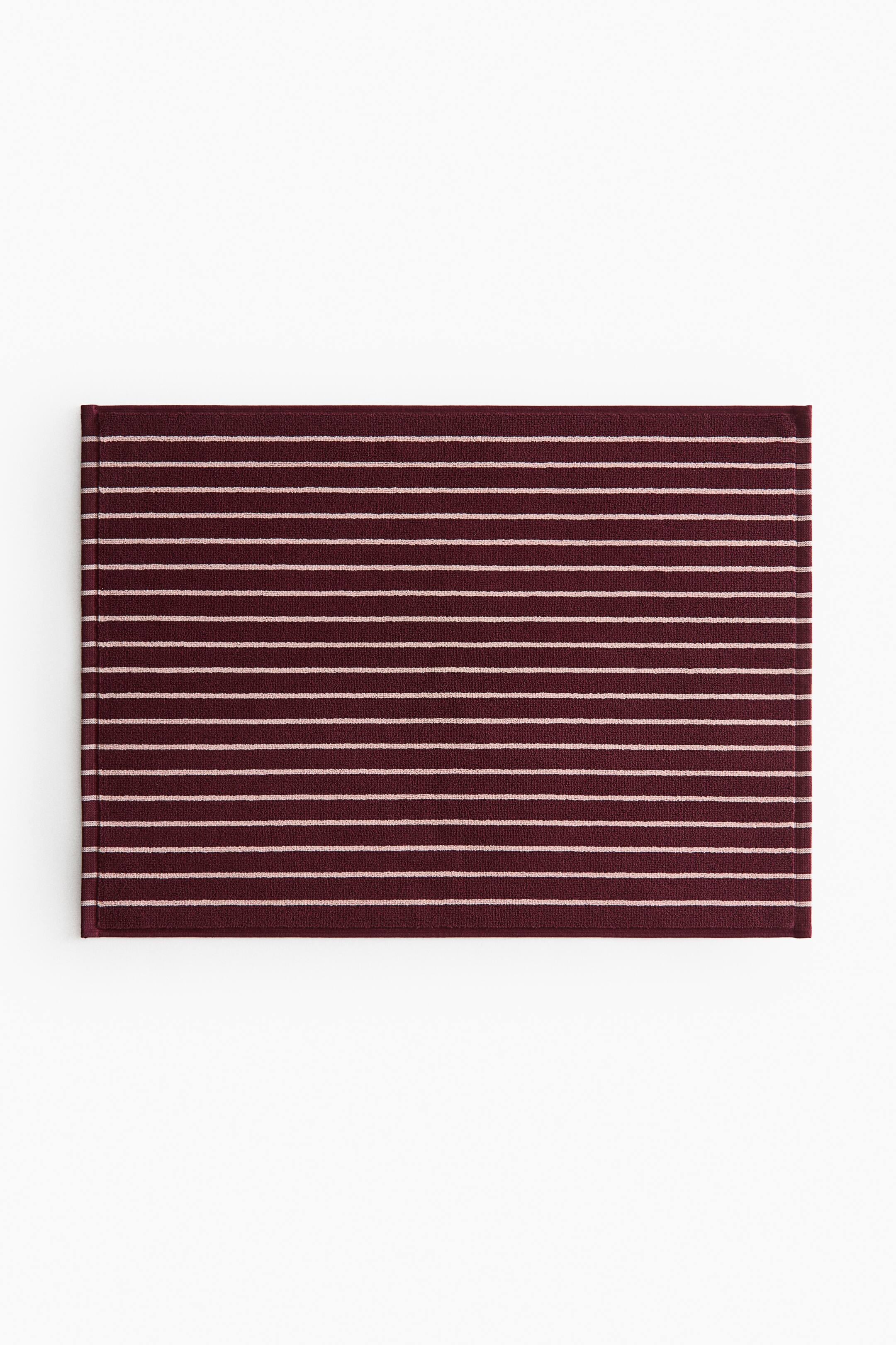View larger image: A rectangular bath mat lies flat, showing a striped pattern. It features horizontal maroon and light pink stripes on a textured surface, with woven trim finishing the edges.
