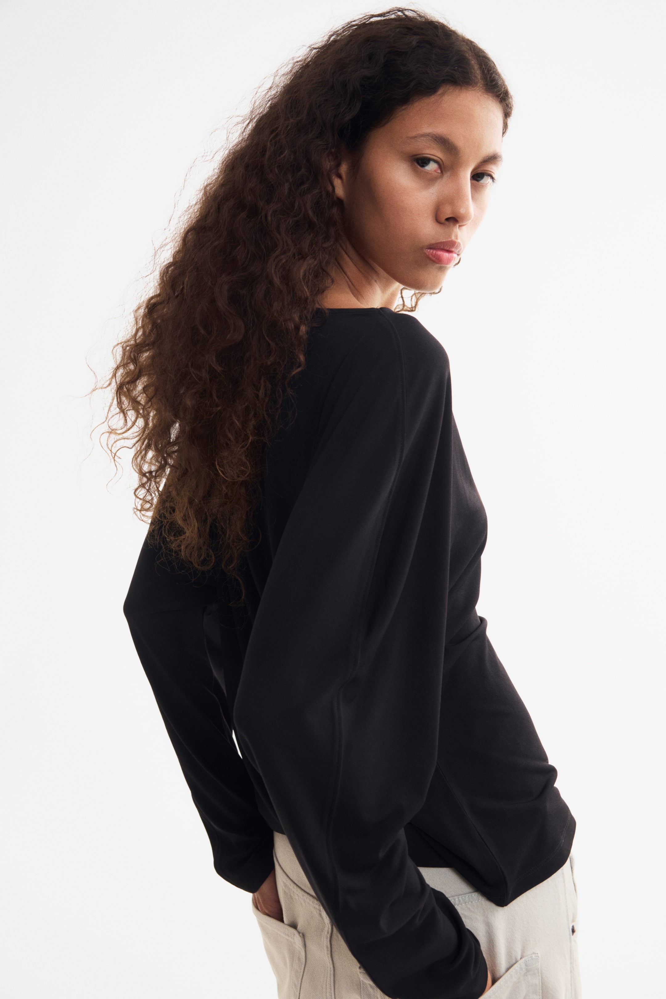 View larger image: A black top in soft stretch jersey, featuring a delicate boat neckline, visible seams down the front and back, long dolman sleeves, and a straight-cut hem, is worn by the model. The model is placing her hands in the pockets of light-coloured trousers.
