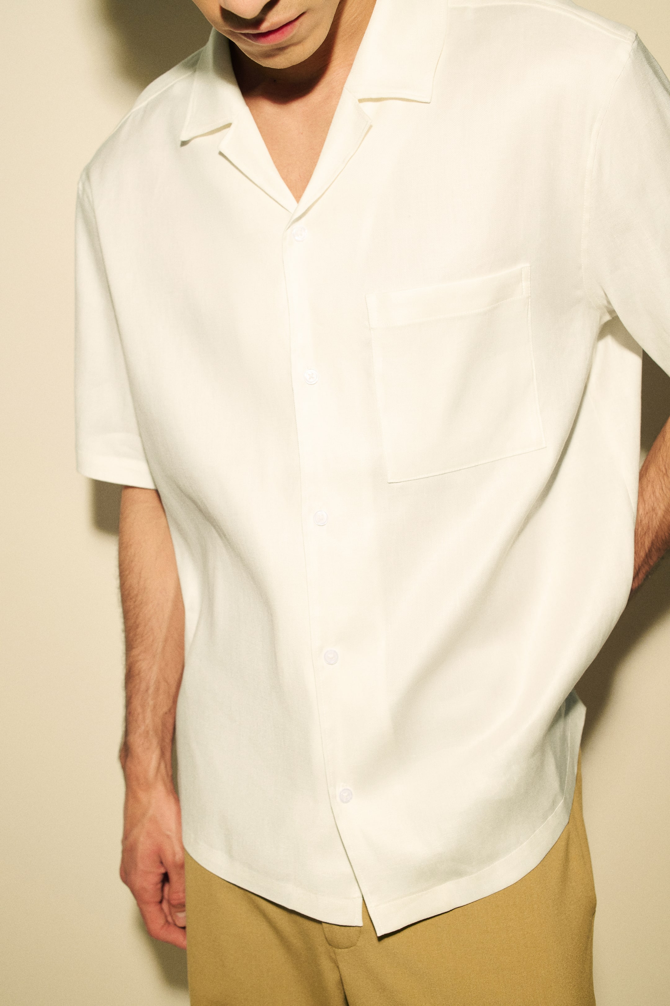 View larger image: The cream short-sleeve shirt is displayed from a front-left angle, showcasing a Cuban collar and a full front button placket with white buttons. A patch pocket is visible on the left chest, and the shirt is designed with a relaxed fit and a curved hem.