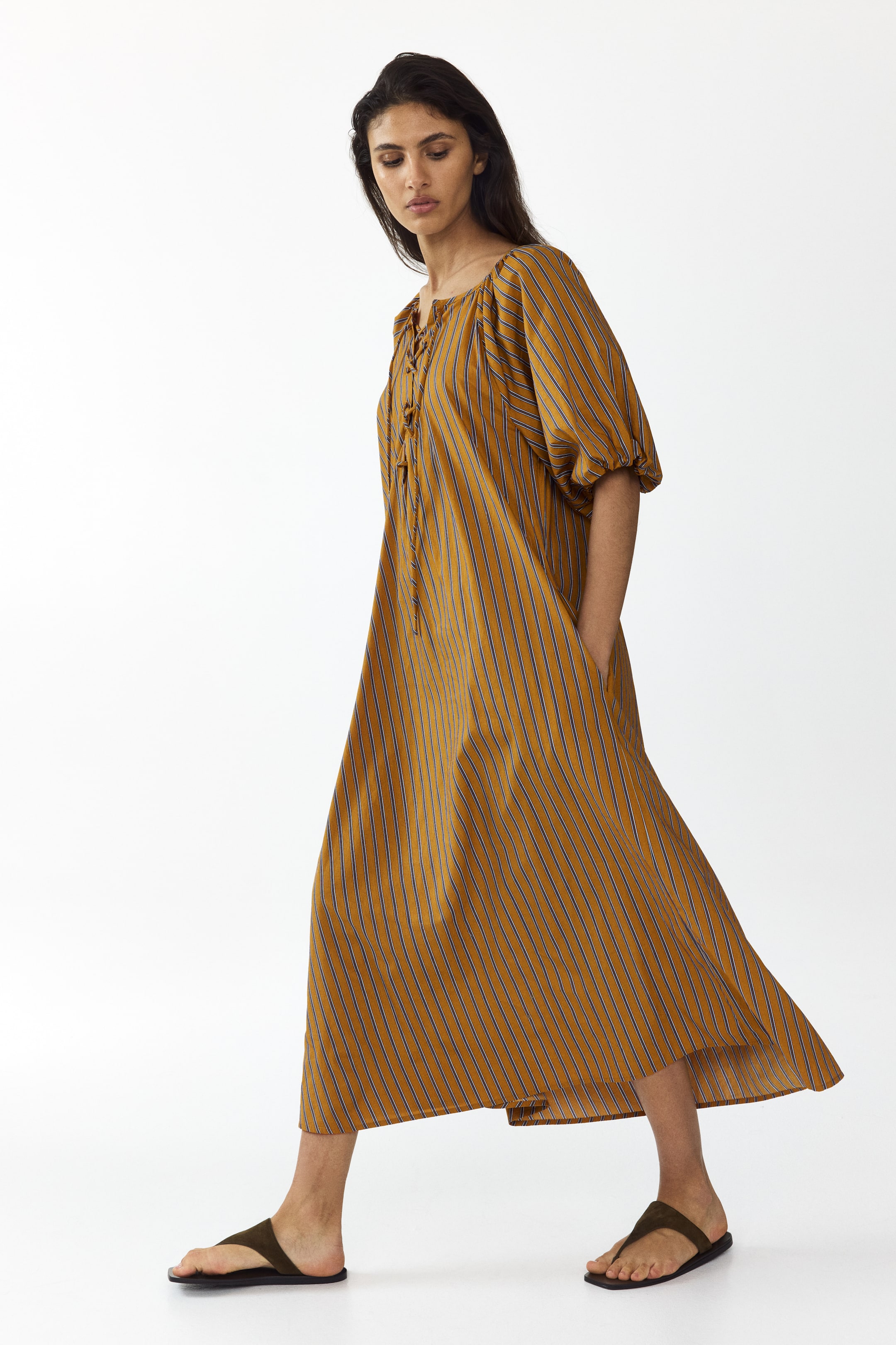 Ingrandisci l'immagine: Un abito maxi giallo senape con sottili righe verticali blu scuro presenta una silhouette ampia e fluente, caratterizzato da uno scollo arricciato con dettaglio allacciato e maniche a sbuffo fino al gomito.