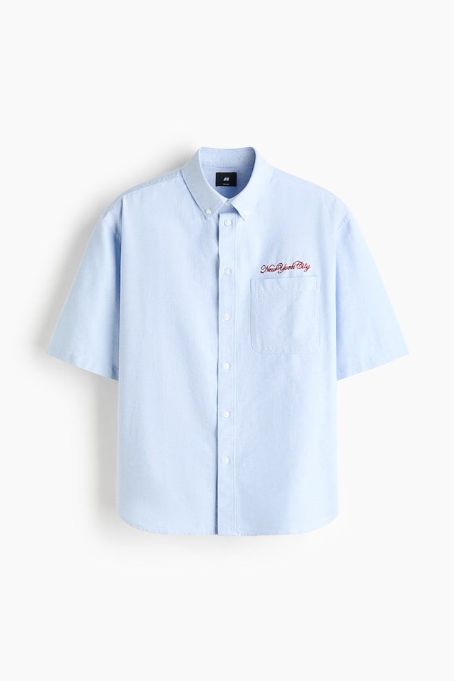 The light blue shirt is shown from the front, featuring a button-down collar and a traditional button-up front. It has short sleeves, an open chest pocket on the left side with red embroidered "New York City" text, and a gently rounded hem, offering a relaxed fit. - 1