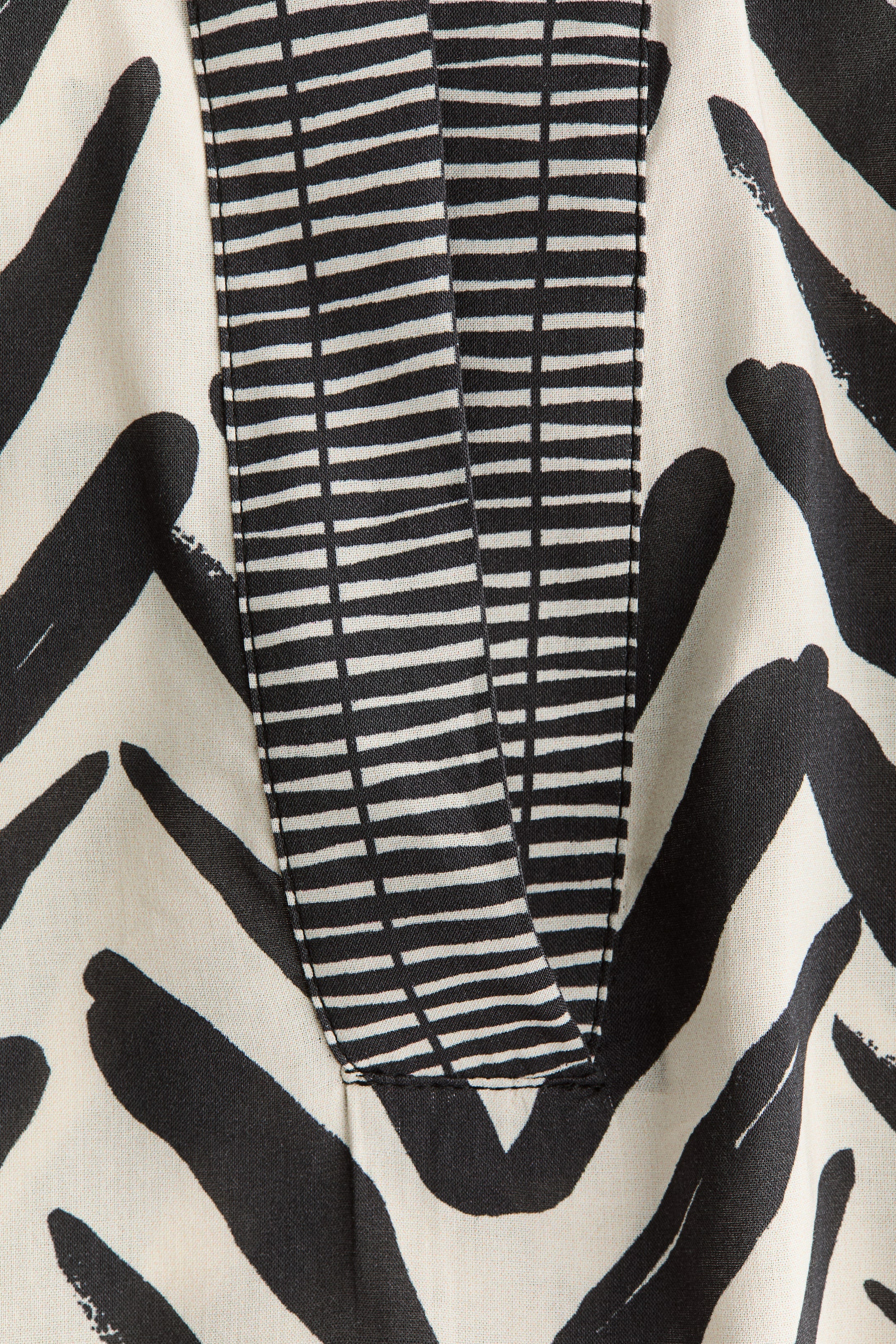 View larger image: This garment showcases an off-white fabric adorned with abstract black brushstroke patterns. A contrasting black placket, featuring thin white horizontal stripes, runs vertically down the front, culminating in a V-shape with visible stitching.