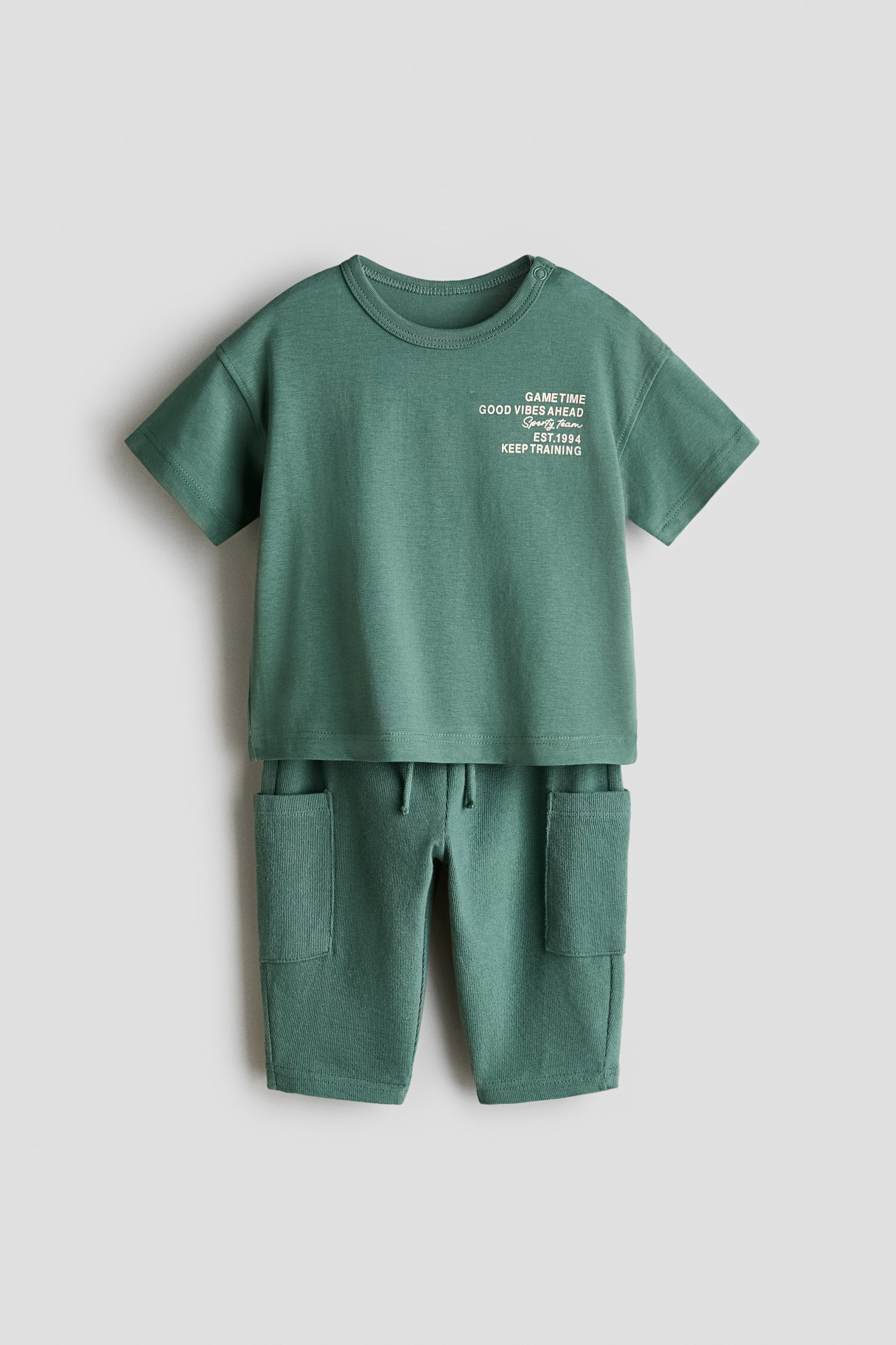 View larger image: A green two-piece set, shown facing front, features a short-sleeved T-shirt with a round neck, snap buttons on the left shoulder and white text on the front reading 'GAMETIME GOOD VIBES AHEAD Sporty Team EST.1994 KEEP TRAINING'. The set also includes matching knee-length shorts with an elasticated drawstring waist and two large front patch pockets, all in a textured knit fabric.