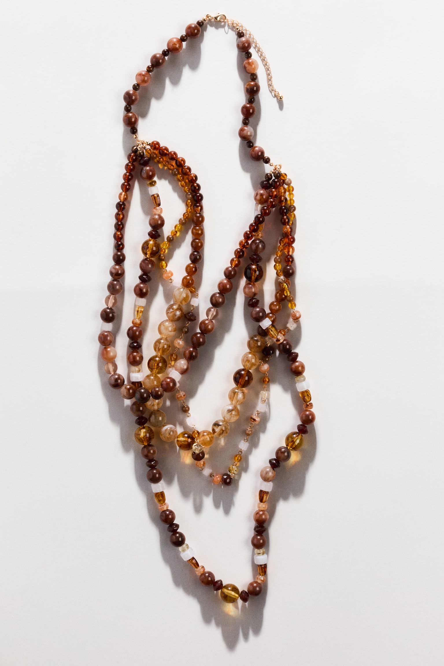 "A multi-strand beaded necklace is showcasing its front, featuring numerous round and rectangular beads in varying shades of brown, amber, and golden yellow. Multiple strands cascade downwards, creating a layered effect, and are connected by a gold-toned adjustable chain at the top." - 1