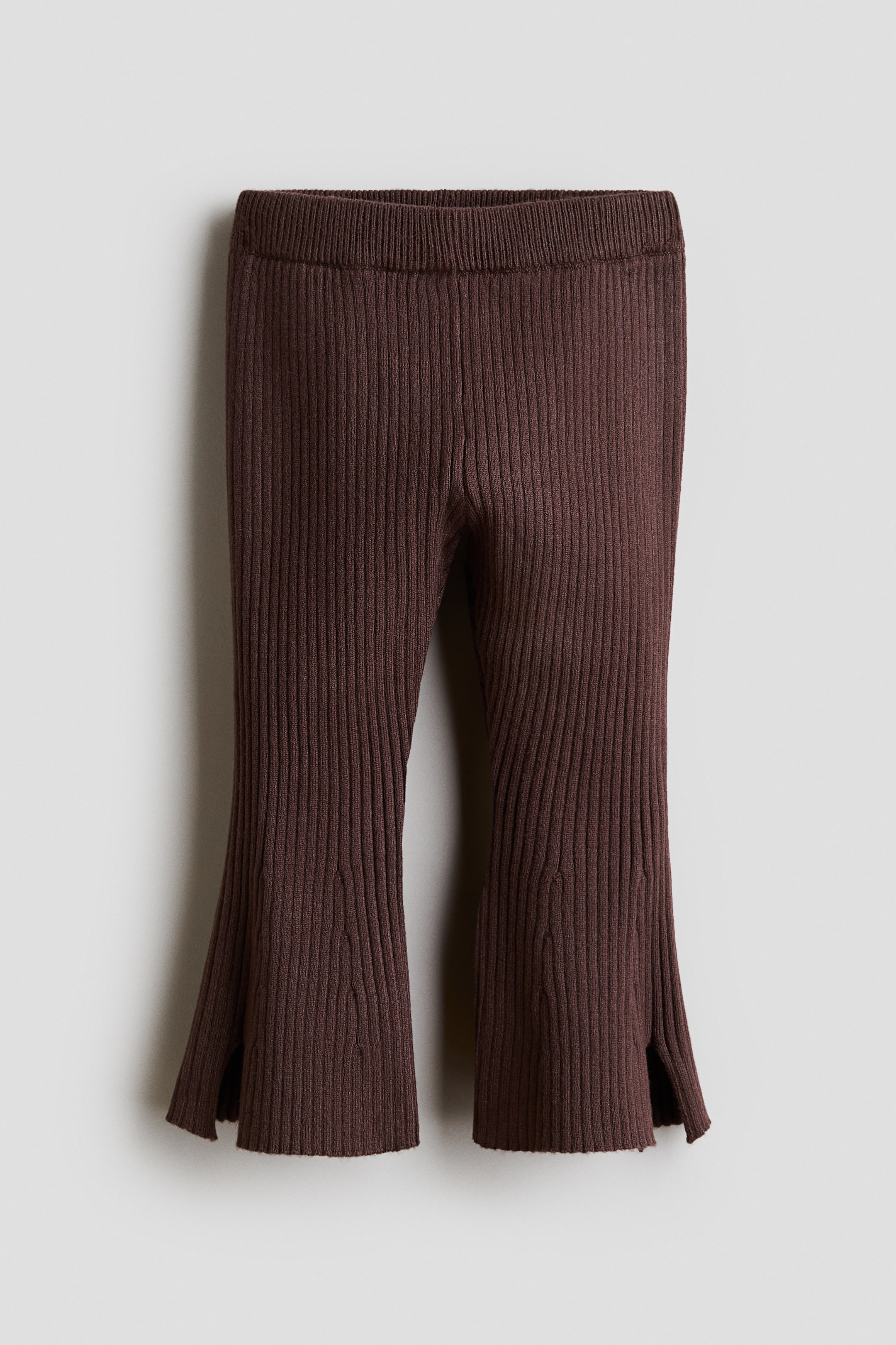 View larger image: A pair of dark brown rib-knit pants is shown from the front, highlighting a vertical ribbed texture and a thick elasticated waistband. The legs flare towards the hem, which features small slits on the outer sides.