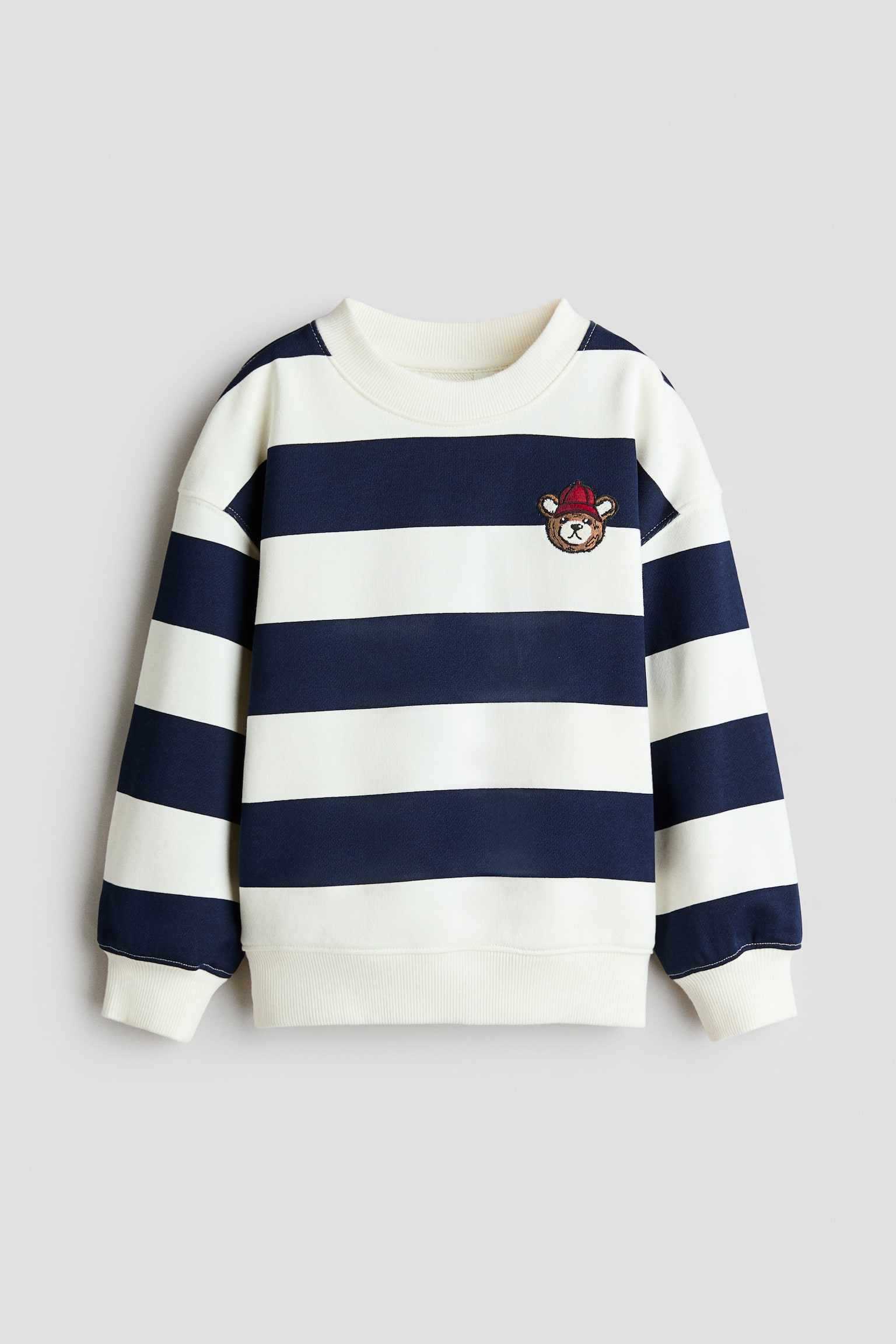 A loose-fit sweatshirt is facing front, displaying horizontal navy blue and off-white stripes with dropped shoulders and a round, ribbed neckline. The garment features a small embroidered bear wearing a red cap on the upper chest, finished with off-white ribbed cuffs and a matching ribbed hem.