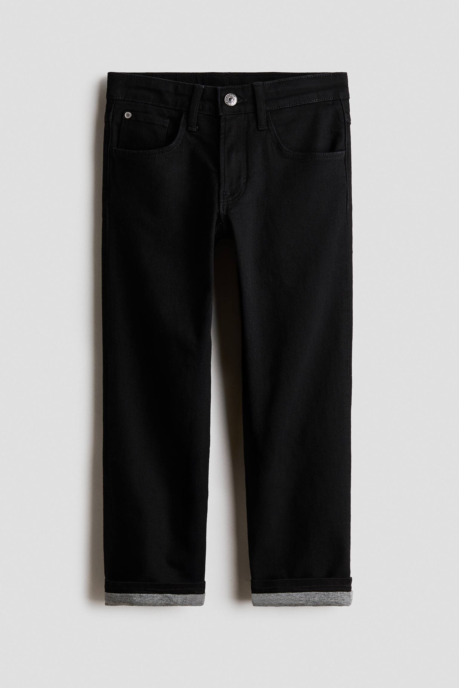 Ett par svarta jeans med raka ben visas framifrÃ¥n och har fem fickor, hÃ¤llor fÃ¶r bÃ¤lte och en silverfÃ¤rgad knappstÃ¤ngning. De nedvikta bensluten visar ett kontrasterande grÃ¥tt foder.