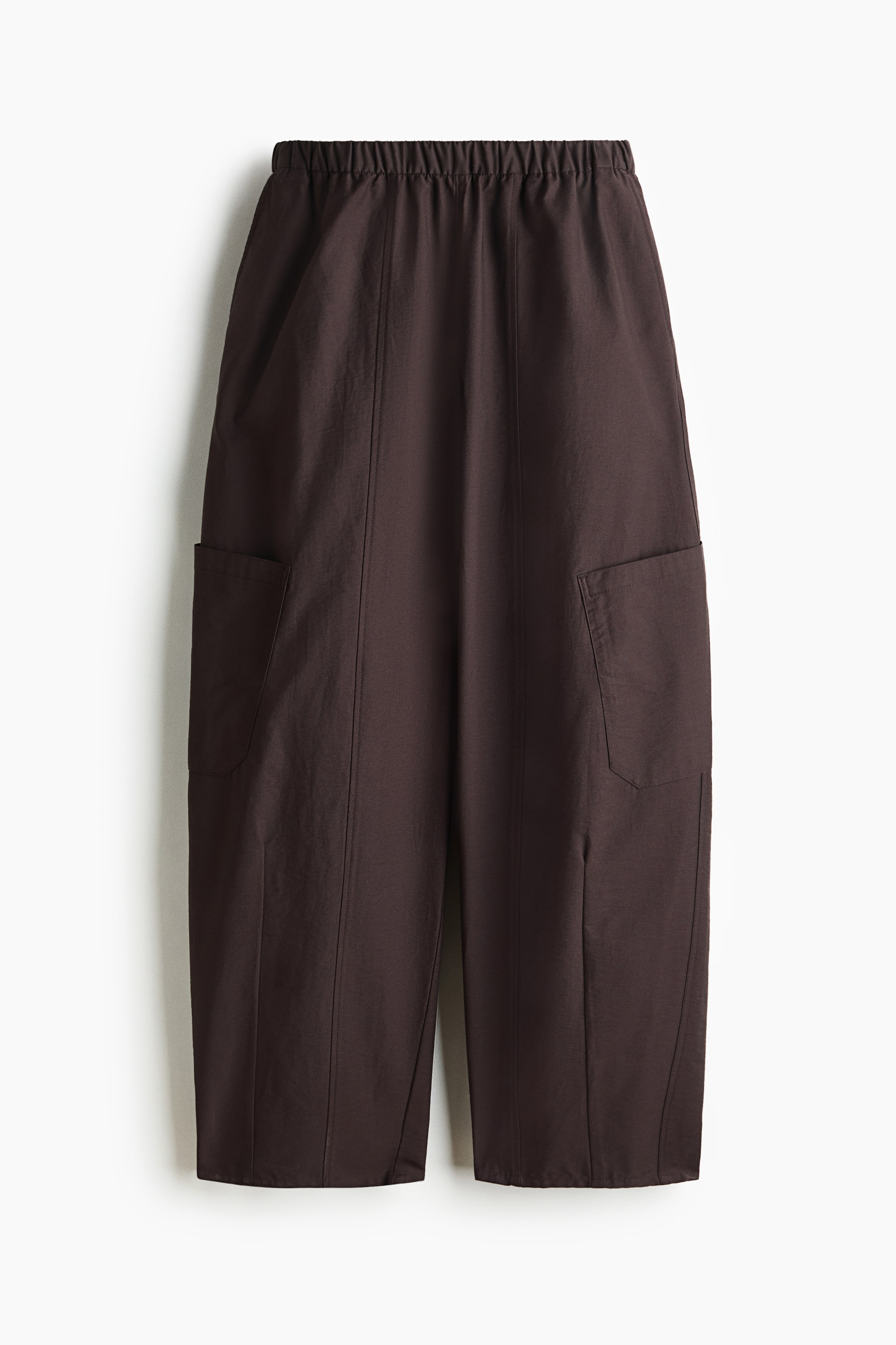 Vezi imaginea mai mare: Pantalonii de culoare maro închis sunt prezentați din față, având o talie elasticizată și picioare largi, scurtate. Două buzunare mari aplicate sunt vizibile în față, câte unul pe fiecare parte, cu cusături verticale care se întind pe fiecare picior.