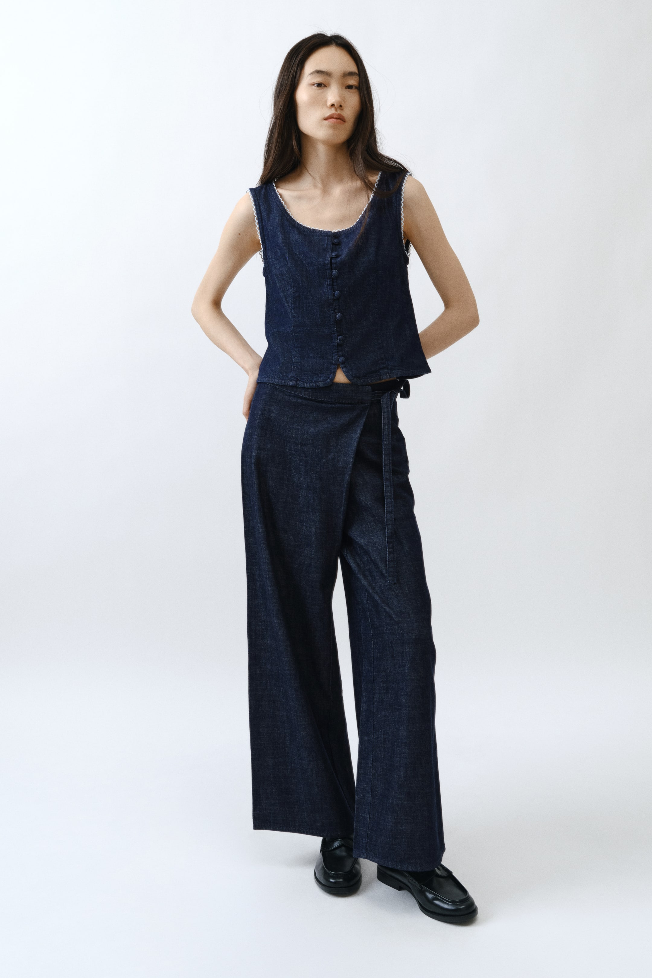 拡大画像を表示: ダークウォッシュデニムのセットアップが紹介されています。ノースリーブのクロップドトップは、スクープネックラインと縁に沿ったホワイトトリムが特徴で、小さなボタンで前面が留められています。ワイドレッグのラップパンツは、ウエストでタイクロージャーによって固定されています。