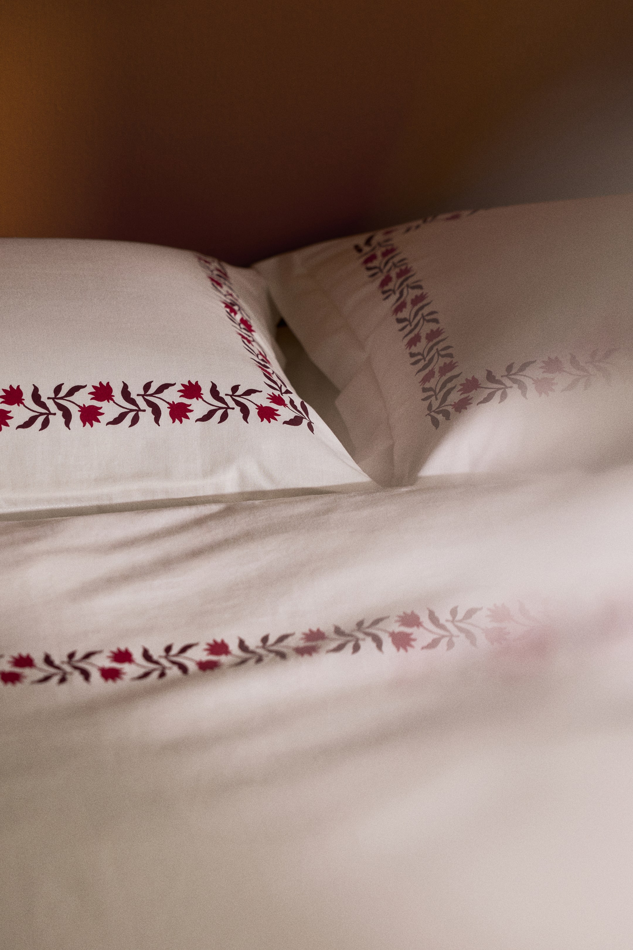 View larger image: A white cotton duvet cover set features a repeating red and dark purple floral vine border along the edges of the duvet cover and pillowcases. Two matching pillowcases are placed behind the folded duvet cover, showcasing the coordinated printed design.