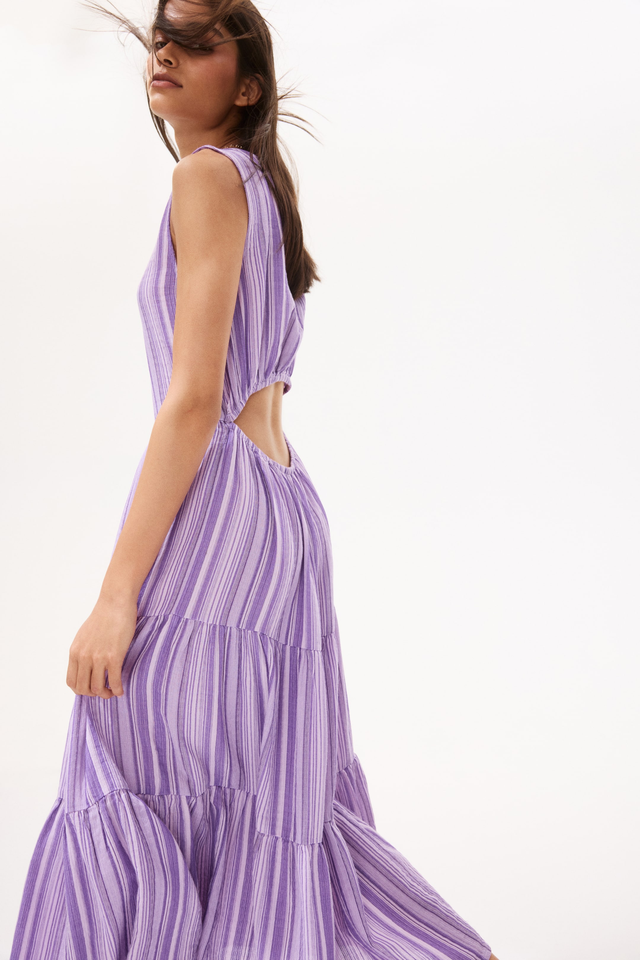 View larger image: "A purple and white striped maxi dress showcases diverse vertical stripes, viewed from a side and back perspective. Crafted with a sleeveless bodice, it features a gathered waist with side cut-outs and a flowing tiered skirt."
