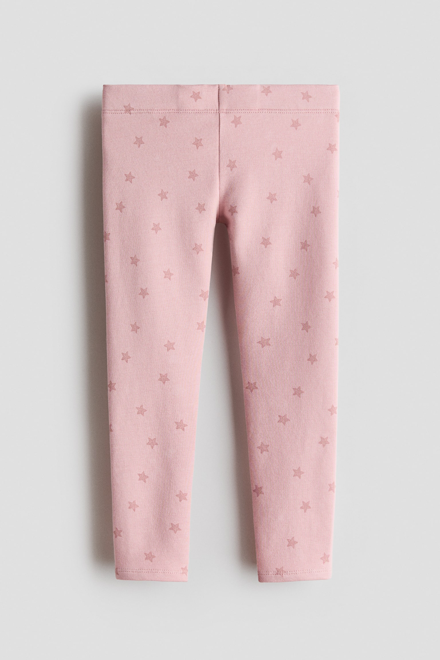 A front view of pink cotton jersey leggings displaying an all-over pattern of small, muted pink stars. The leggings feature an elasticized waistband and slim legs tapering to the ankles.