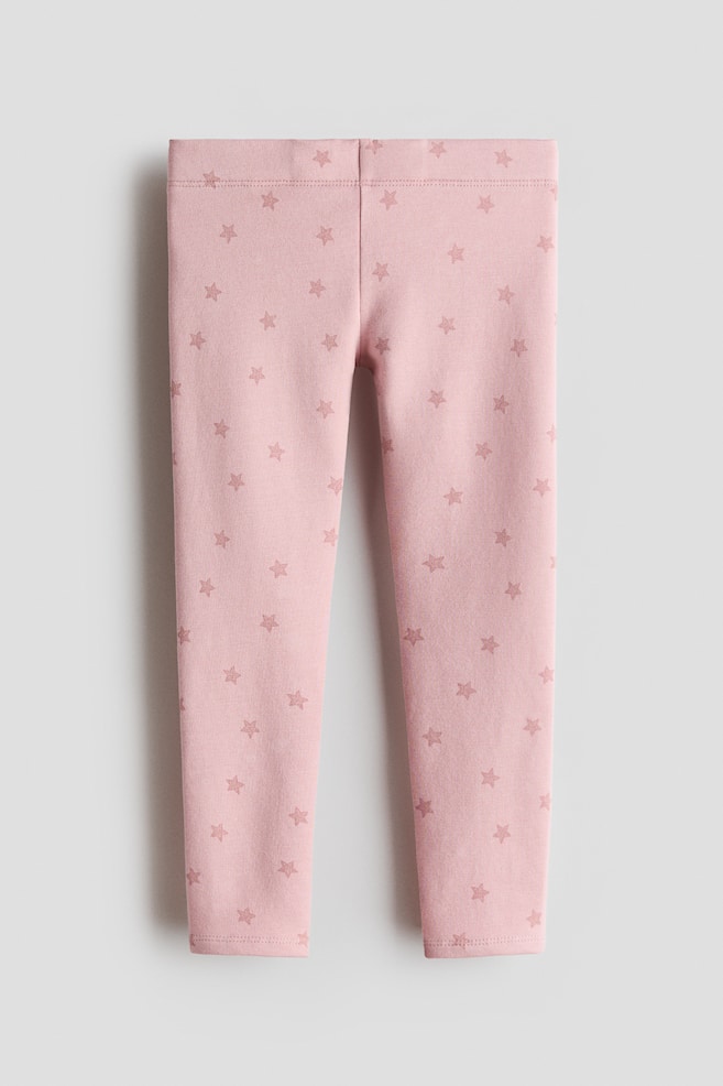 A front view of pink cotton jersey leggings showing an all-over pattern of small, muted pink stars. The leggings feature an elasticated waistband and slim legs tapering to the ankles. - 1