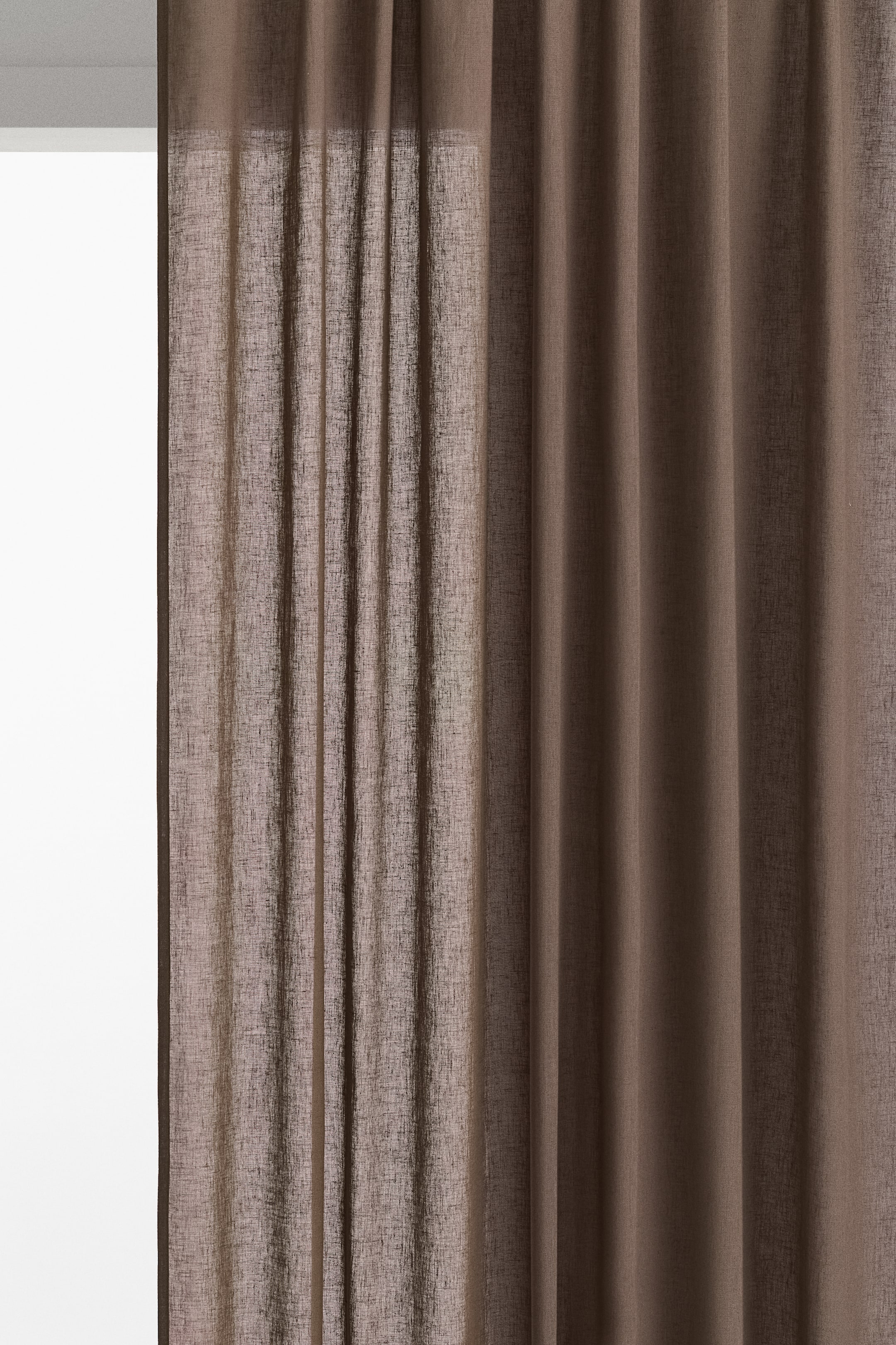 Visa större bild: Ett par bruna gardinlängder visar en texturerad linne- och bomullsväv. En gardin är delvis dragen åt sidan, vilket låter ljuset sippra igenom, medan den andra hänger i mjuka veck, båda med ett multifunktionellt huvud med synliga små veck längs toppen.