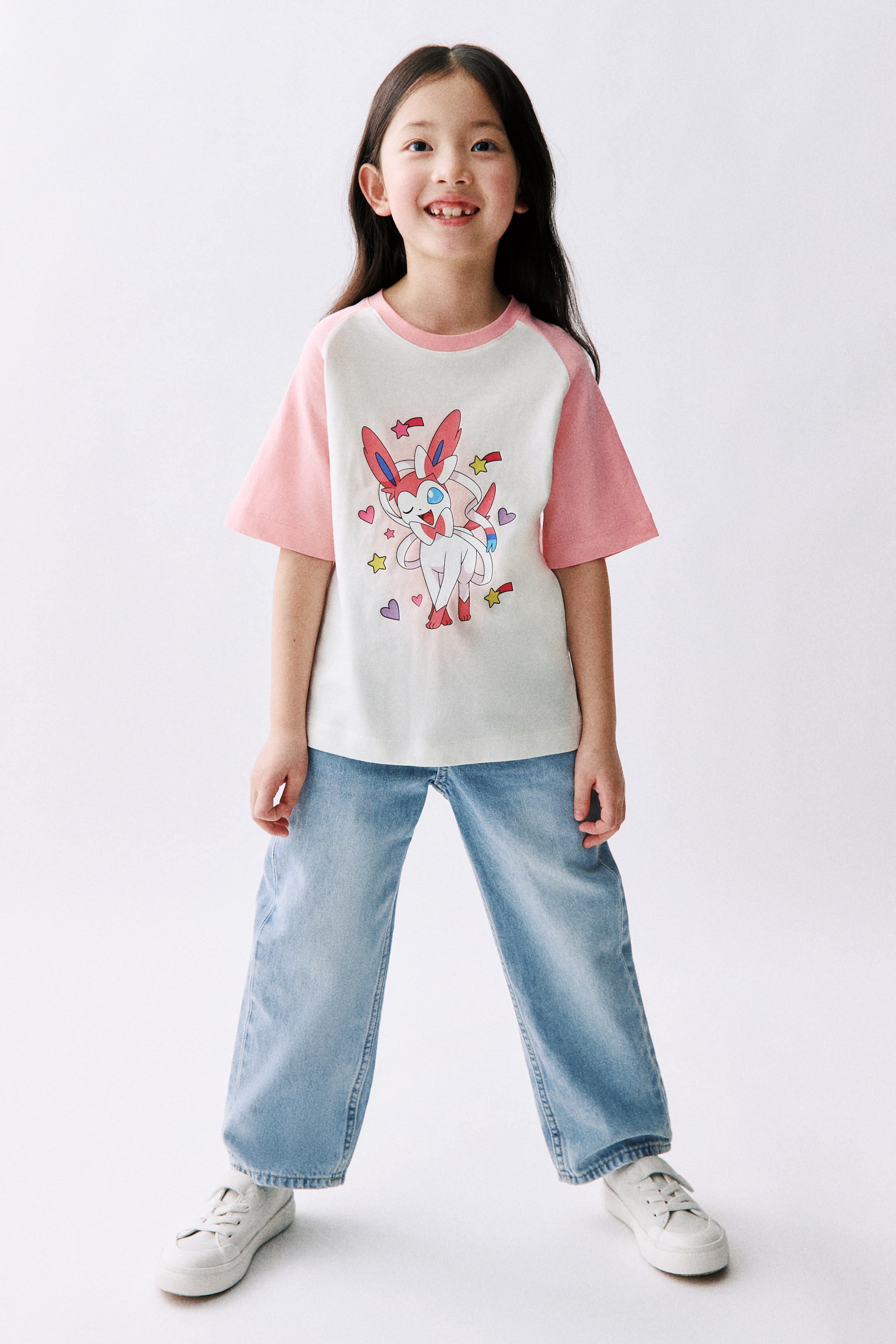 Xem ảnh lớn hơn: Áo thun có thân màu kem với tay áo raglan ngắn màu hồng. Một hình ảnh đầy màu sắc của Sylveon, được bao quanh bởi các ngôi sao và trái tim, được in ở phía trước.