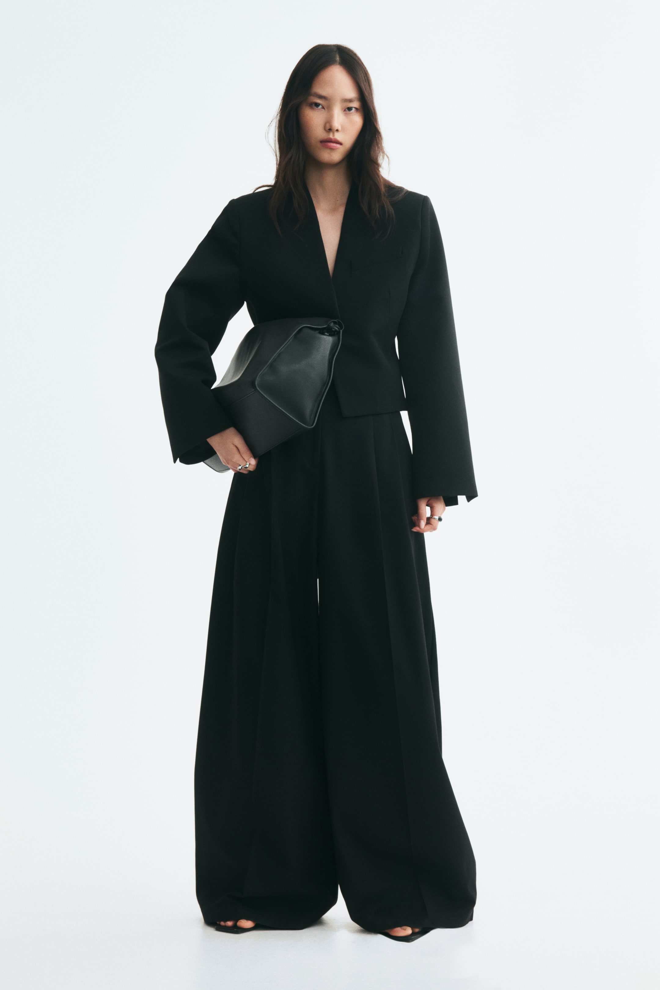 Größeres Bild anzeigen: Locker geschnittene schwarze Hose aus Twill mit voluminösem Barrel-Leg und Bundfalten, getragen vom Model. Die Hose ist mit einem passenden verkürzten schwarzen Blazer kombiniert, dazu eine große schwarze Ledertasche, die unter dem Arm des Models getragen wird.