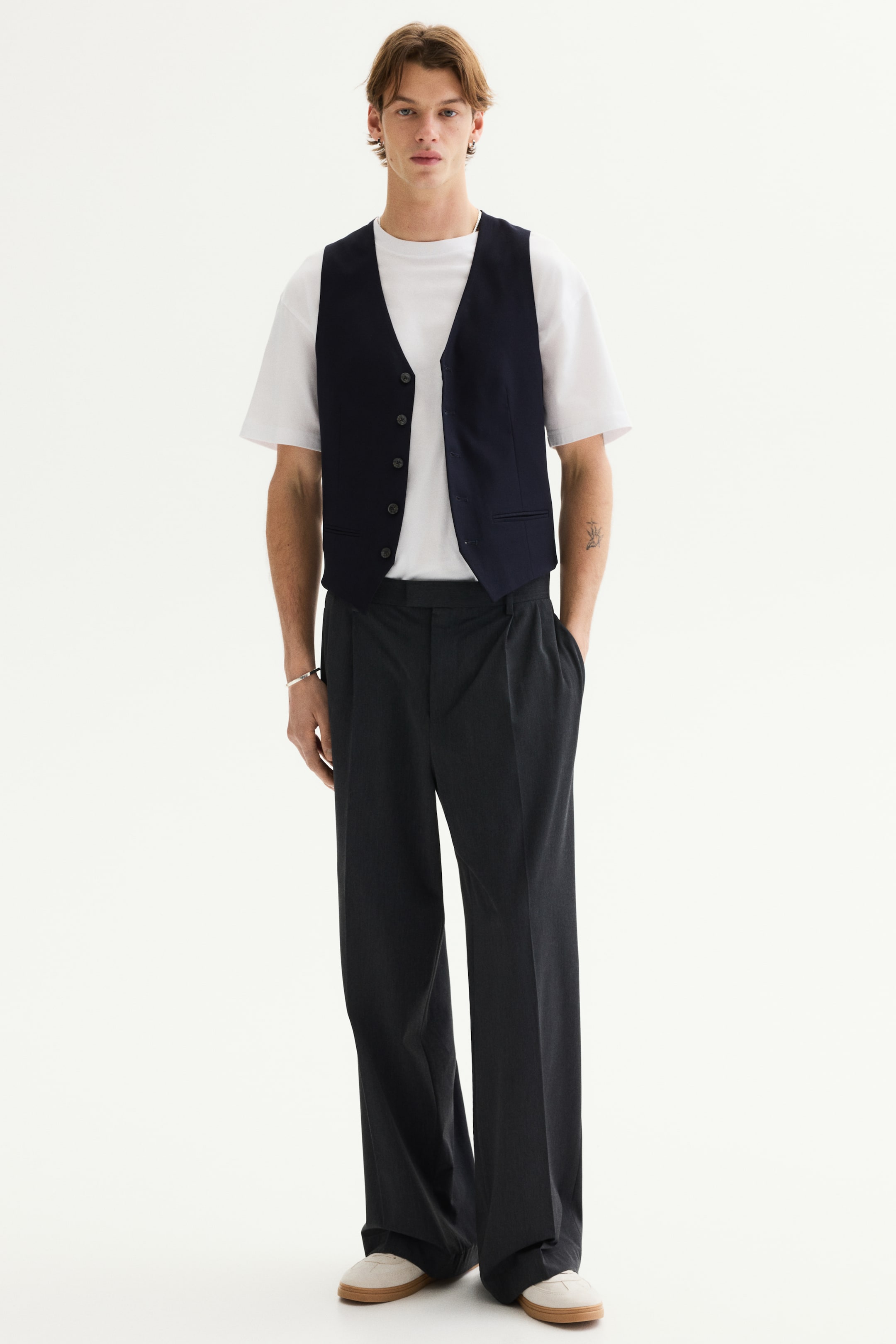 Ver imagen más grande: Un chaleco de botones al frente en color azul marino oscuro está sobre una camiseta blanca de cuello redondo, y presenta un escote en V con seis botones visibles. Unos pantalones de pierna ancha y pliegues en color gris oscuro completan el conjunto, y se llevan con zapatillas de color claro.