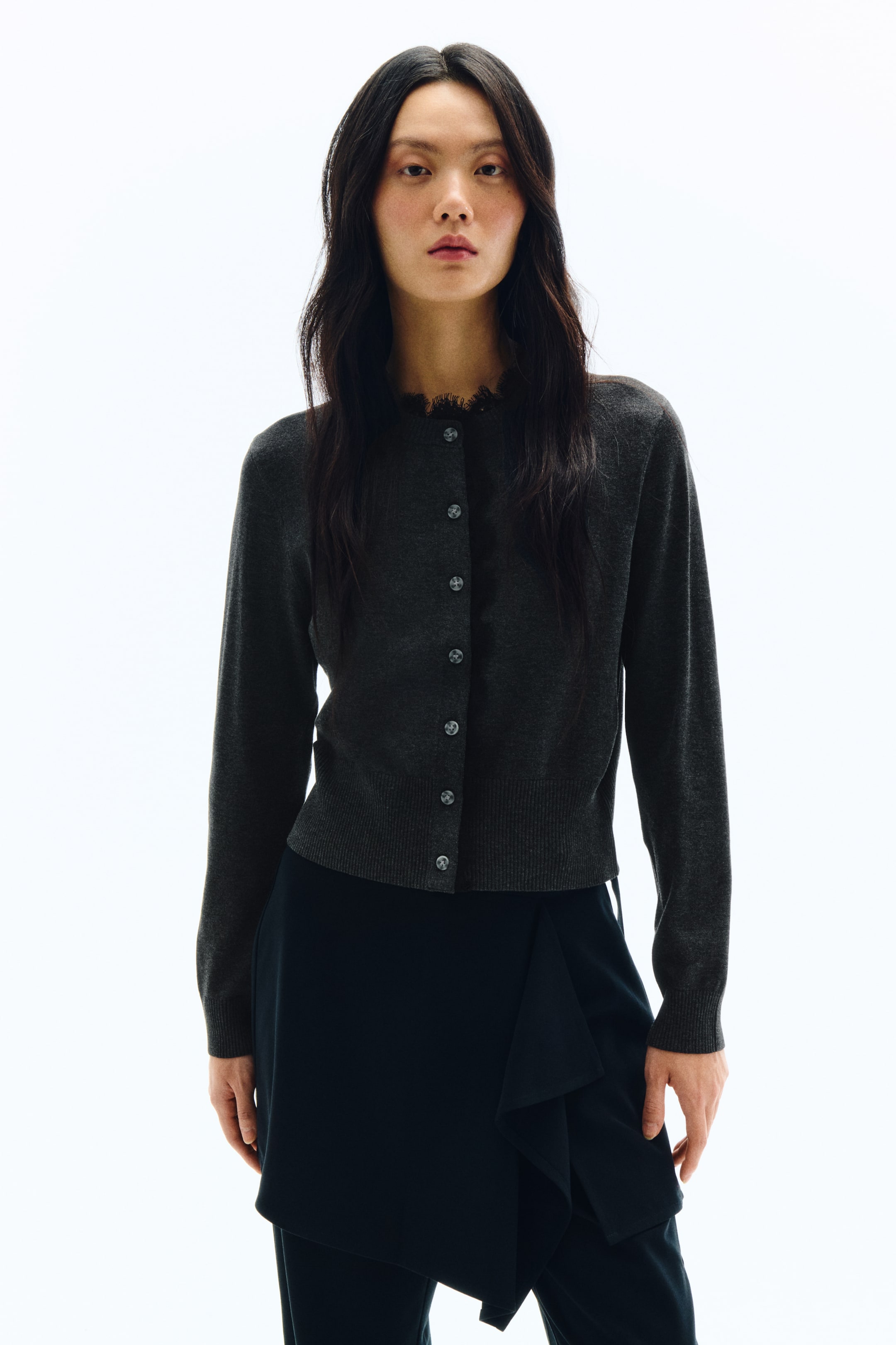 拡大画像を表示: ダークグレーのカーディガンが正面向きで写っており、フリンジ付きトリムのクルーネックデザインになっています。フロントには中央に並んだダークカラーのボタンが7つ配され、袖口と裾はリブ仕様です。