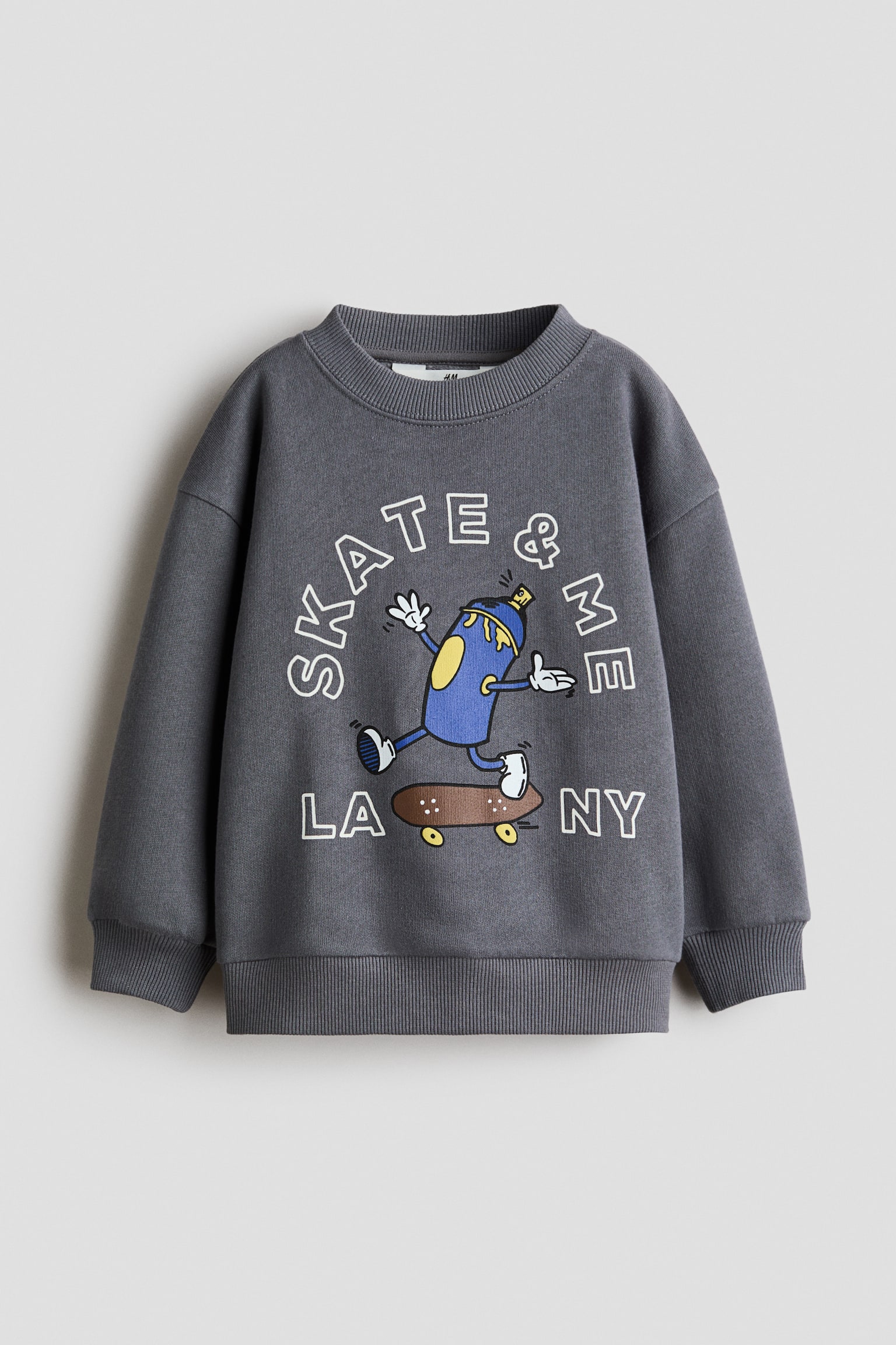 A dark grey sweatshirt is shown from the front, featuring a round ribbed neckline, dropped shoulders, and long sleeves with ribbed cuffs. The front displays a colourful graphic print of a cartoon spray can character riding a skateboard, surrounded by the white outlined text âSKATE & MEâ and âLA NYâ. - 1