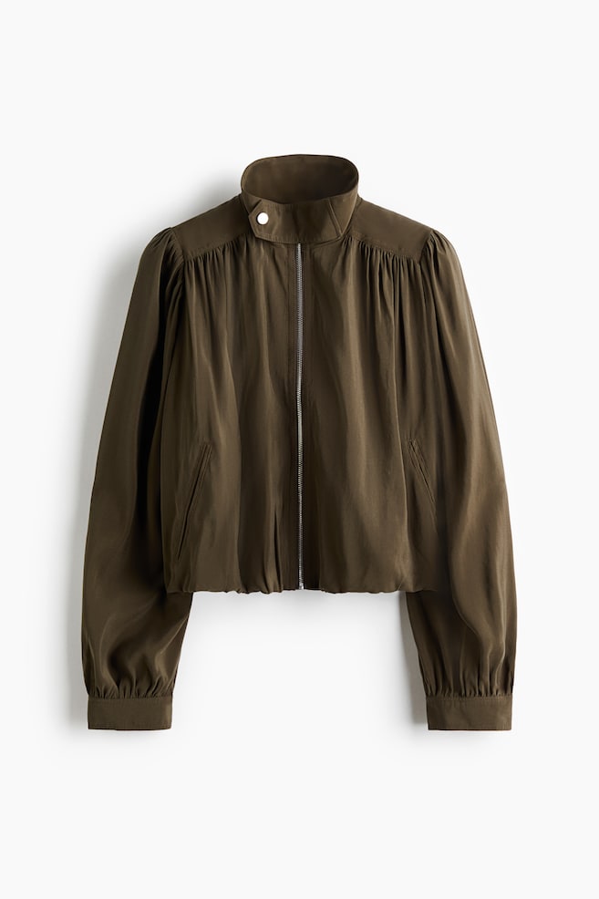 An olive green cropped jacket is presented with a high stand-up collar secured by a snap button and a central silver-toned zip. It showcases gathered details at the shoulders and above the cuff bands, along with slanted welt pockets and an elasticated hem. - 1