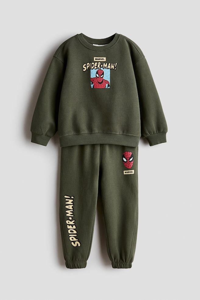 Un chándal infantil de dos piezas en verde oliva se muestra de frente, compuesto por una sudadera de manga larga y un pantalón jogger a juego. La sudadera presenta un estampado gráfico central de Spider-Man con el texto "SPIDER-MAN!", rematado con cuello redondo, puños y bajo de canalé. El jogger muestra un estampado de la máscara de Spider-Man en la cadera izquierda y el texto vertical amarillo "SPIDER-MAN!" en la parte inferior de la pierna derecha, con bajos elásticos. - 1