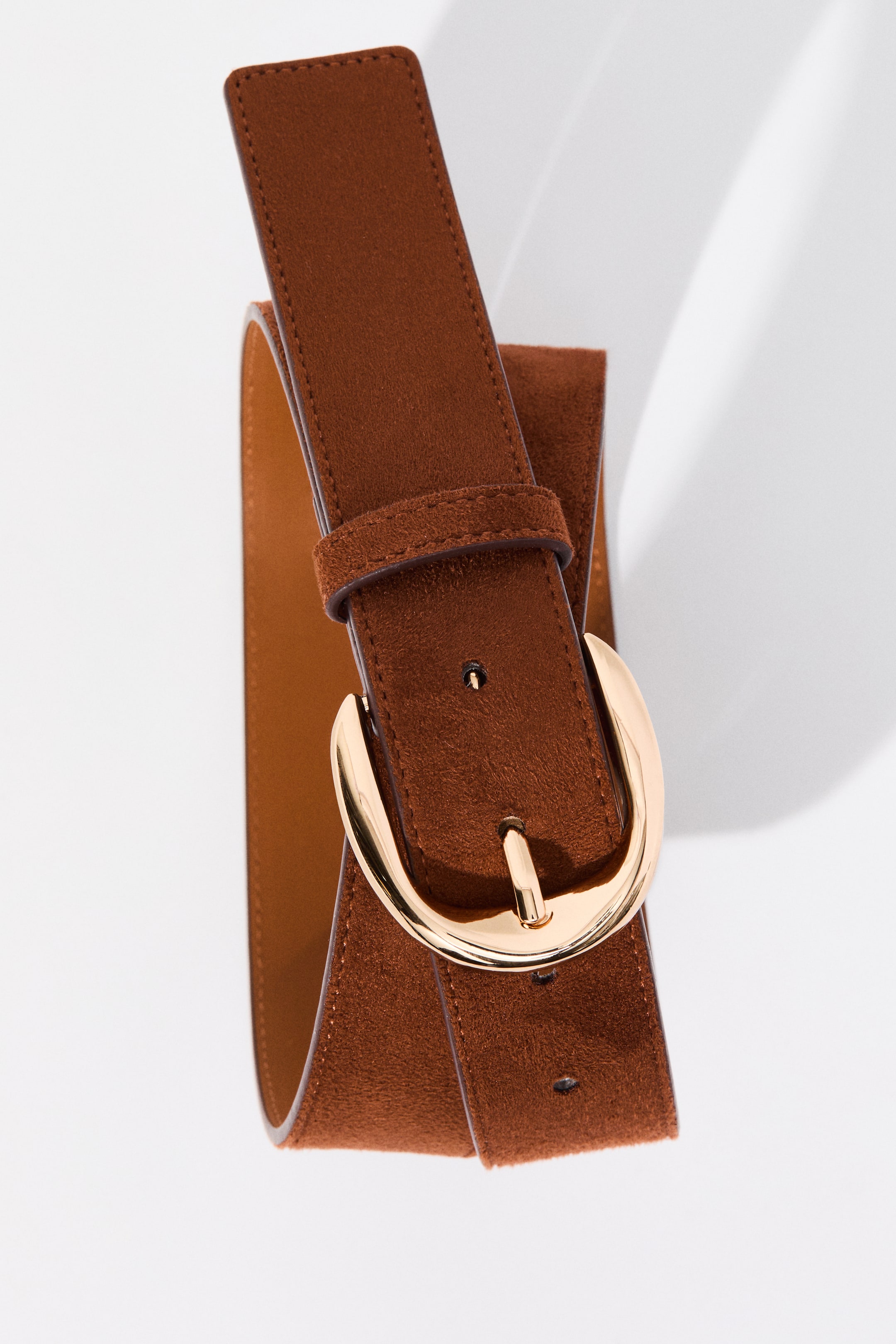 View larger image: A brown suede belt coils elegantly, showcasing its textured surface with a smoother underside. It features a gold-toned D-shaped buckle, a matching keeper, and visible stitching along its edges.