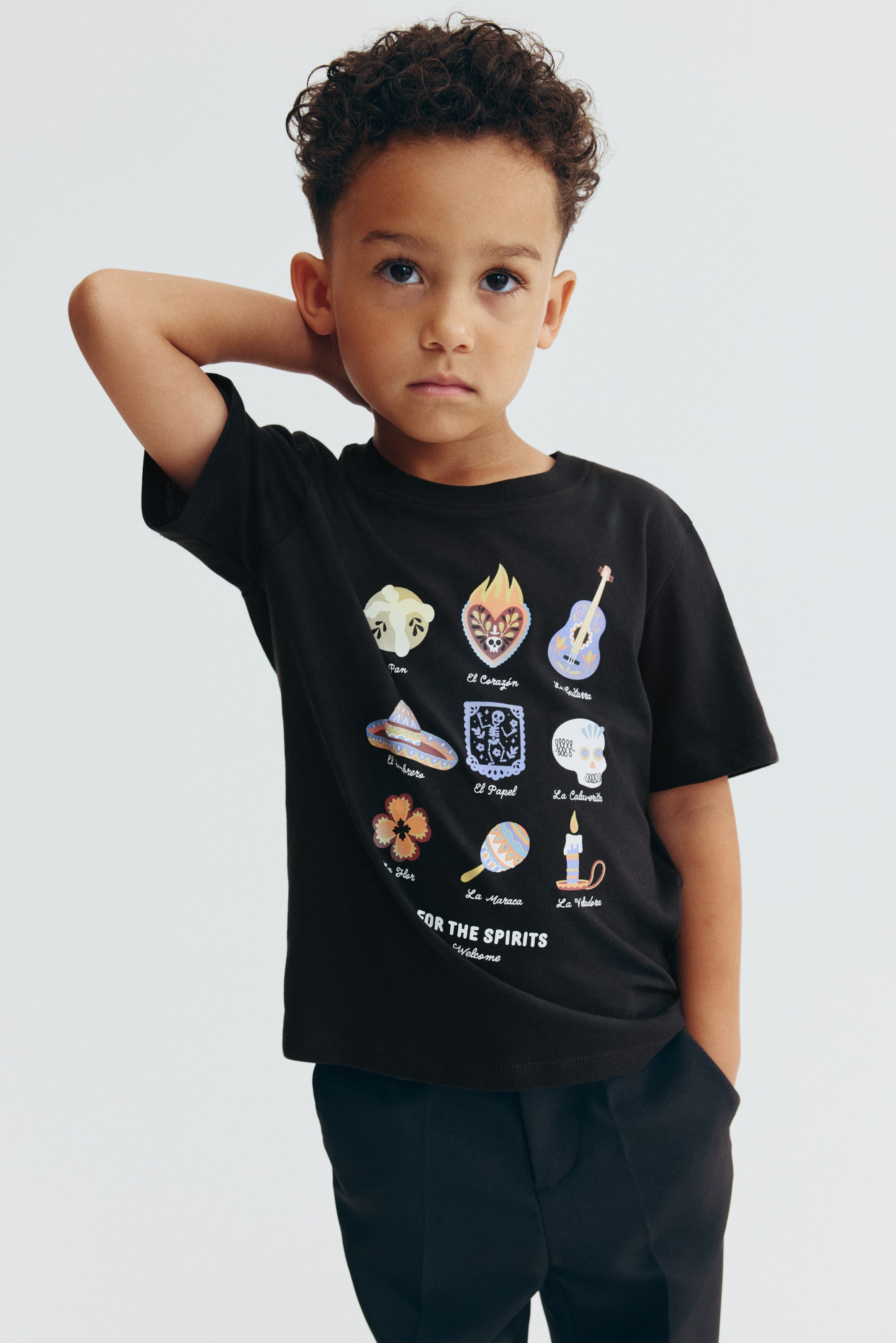 View larger image: A black cotton jersey T-shirt is shown from the front, featuring a ribbed crew neckline, dropped shoulders, and a relaxed fit. The front displays a colorful graphic print of nine illustrated icons arranged in a grid with corresponding text labels, styled with black trousers.