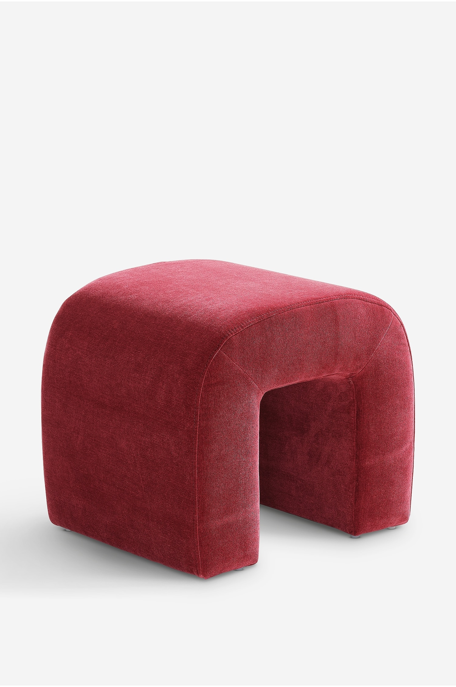 Un pouf a forma di arco, rivestito in velluto rosso, Ã¨ posizionato ad angolazione tre quarti, evidenziando il suo design a U rovesciata. Due libri e un vaso metallico argentato con una pianta verde sono appoggiati sulla sua superficie superiore arrotondata.
