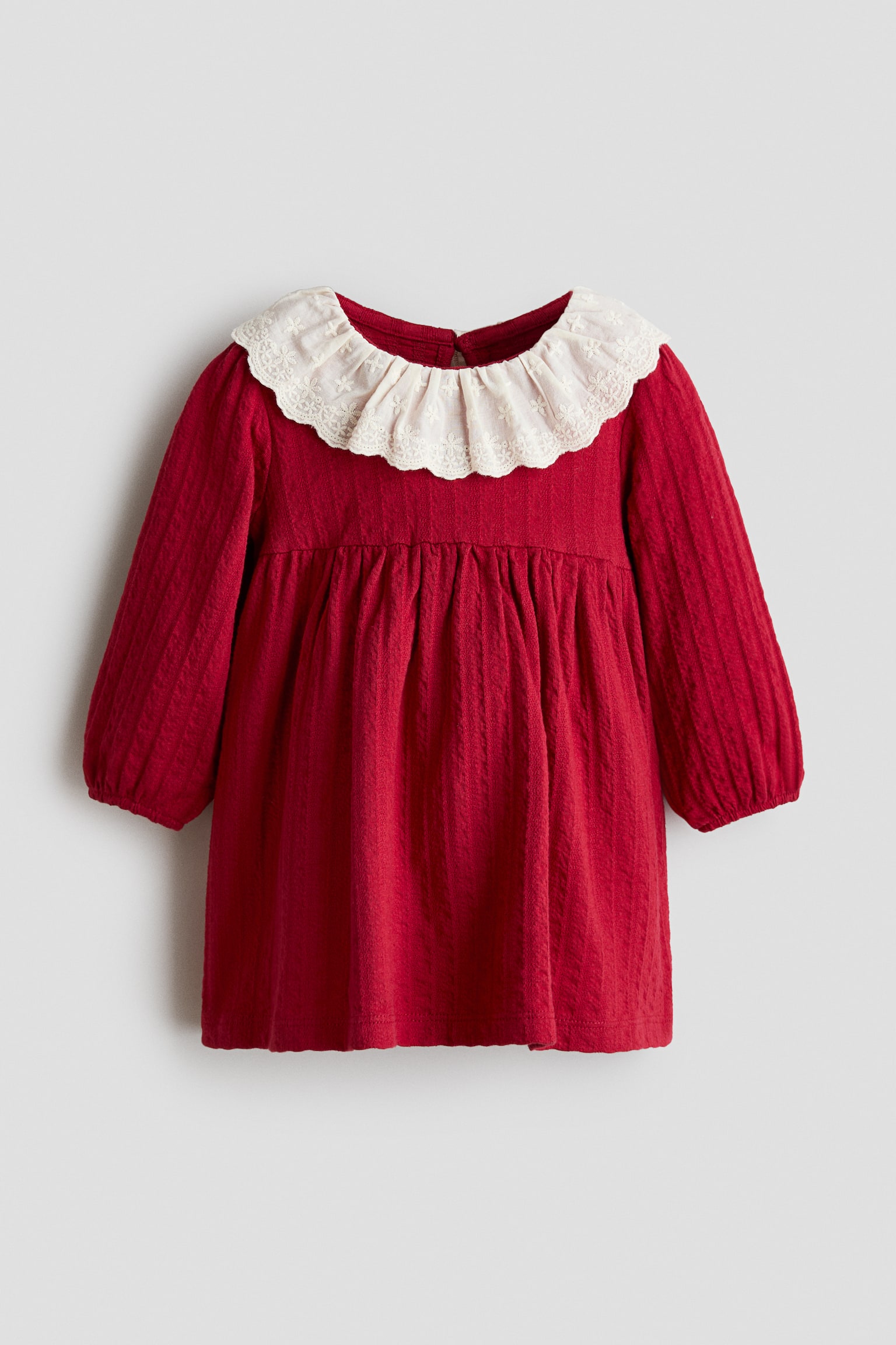 Une robe rouge en jersey de coton texturÃ© est prÃ©sentÃ©e de face, avec un large col en dentelle crÃ¨me Ã  volants, ornÃ© de broderies dÃ©licates. La robe montre de longues manches ballon avec poignets Ã©lastiquÃ©s et une jupe Ã©vasÃ©e froncÃ©e Ã  la taille haute.