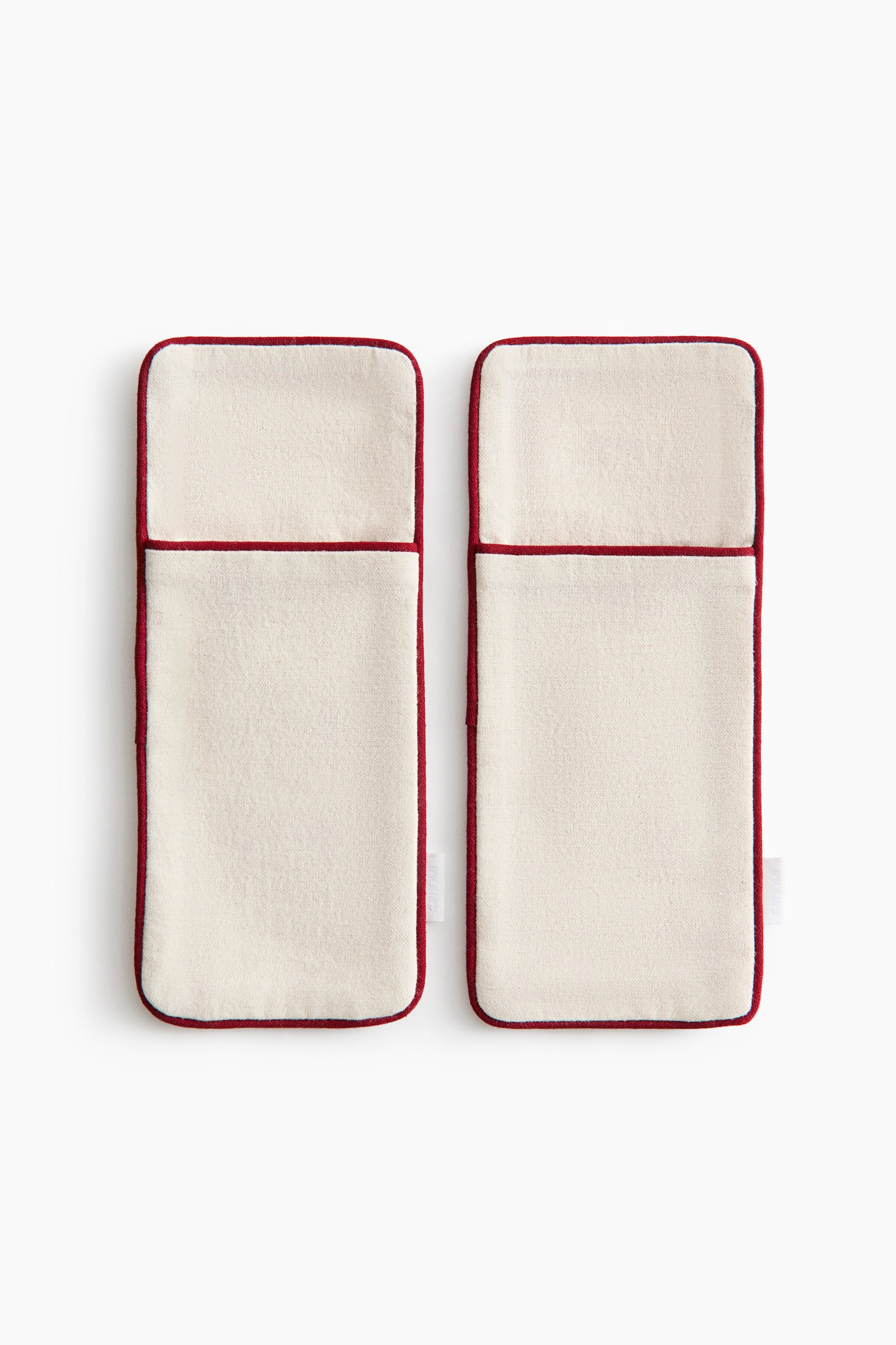 A pair of rectangular, cream-coloured fabric cutlery holders is shown front-on, side by side, showcasing a textured woven material. The holders feature front pockets and contrasting red piped edging along their rounded edges and pocket openings. - 1